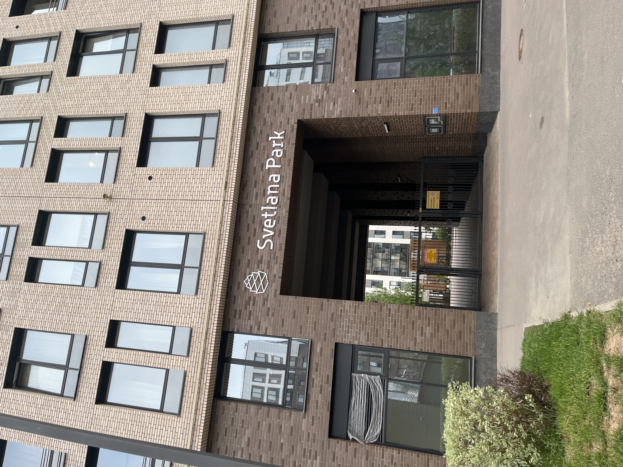 The image shows the entrance to the "Svetlana Park" residential complex, located in Saint Petersburg, Russia. The entrance features a covered walkway leading to a gated area. The building is constructed of beige and brown bricks. The facade displays the name "Svetlana Park" in white lettering and a logo above the entrance. Multiple windows with dark frames are visible on the building's facade. A paved area extends from the building, bordered by grass and bushes.