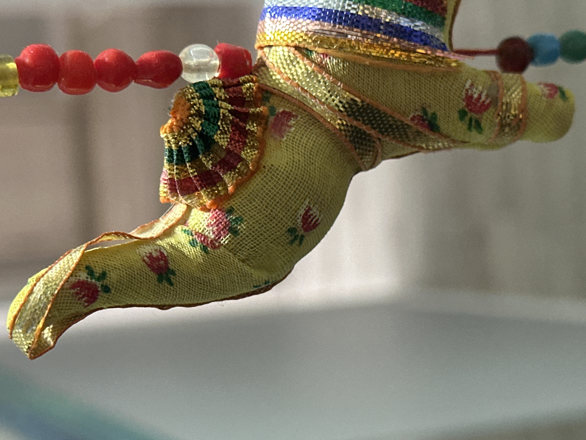 A close-up, horizontal, eye-level shot shows a colorful, fabric-covered elephant ornament. The elephant is yellow with a subtle floral pattern and adorned with metallic gold trim. It has a decorative headdress with red, green, and gold fringed fabric. A strand of red, clear, and yellow beads hangs to the right of the elephant's head, and a string of colorful pom-poms dangles from its lower body. The background is a soft, out-of-focus blur of light gray and white, suggesting an indoor setting or possibly an outdoor shaded area. The lighting is bright, creating highlights on the metallic elements of the ornament.