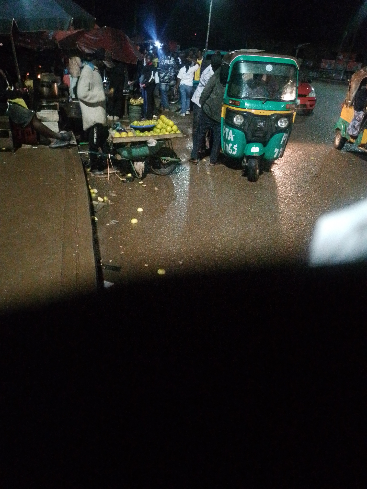 This is a medium shot, slightly high-angle, nighttime street scene in an urban environment. The ground is wet, suggesting recent rain. In the foreground, a person is partially visible on the left, seated with their back to the camera. To the right and slightly further back, a green and yellow three-wheeled auto-rickshaw is parked. It has the license plate "PTA-1765" on its front. Several people are standing around the auto-rickshaw, some appearing to be interacting with it or each other. Further to the right, another auto-rickshaw, predominantly yellow and green, is also visible. In the background, a red car is parked, and there are several market stalls with awnings and lights, indicating a bustling area. A pile of fruit, possibly oranges, is displayed on a cart in front of the auto-rickshaw. The overall atmosphere is that of a busy street market at night, possibly in a developing country, with a wet surface due to rain.