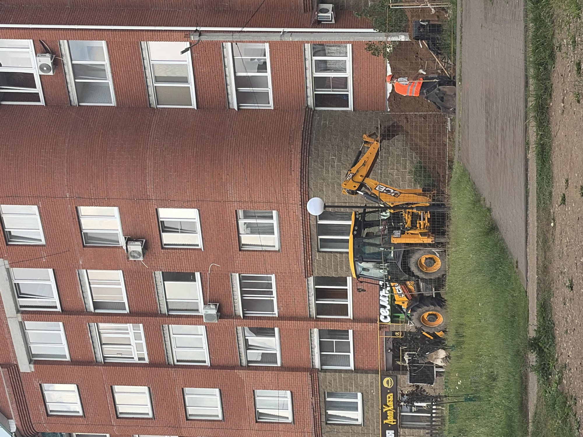 The image shows a residential brick building in the city of Chaikovsky, Russia, with construction work taking place in front of it. An excavator, a construction worker in an orange vest, and a pile of dirt are visible. The building has multiple stories with many windows. The ground in front of the building is a mix of grass and paved areas. The overall scene depicts an active construction or repair site.