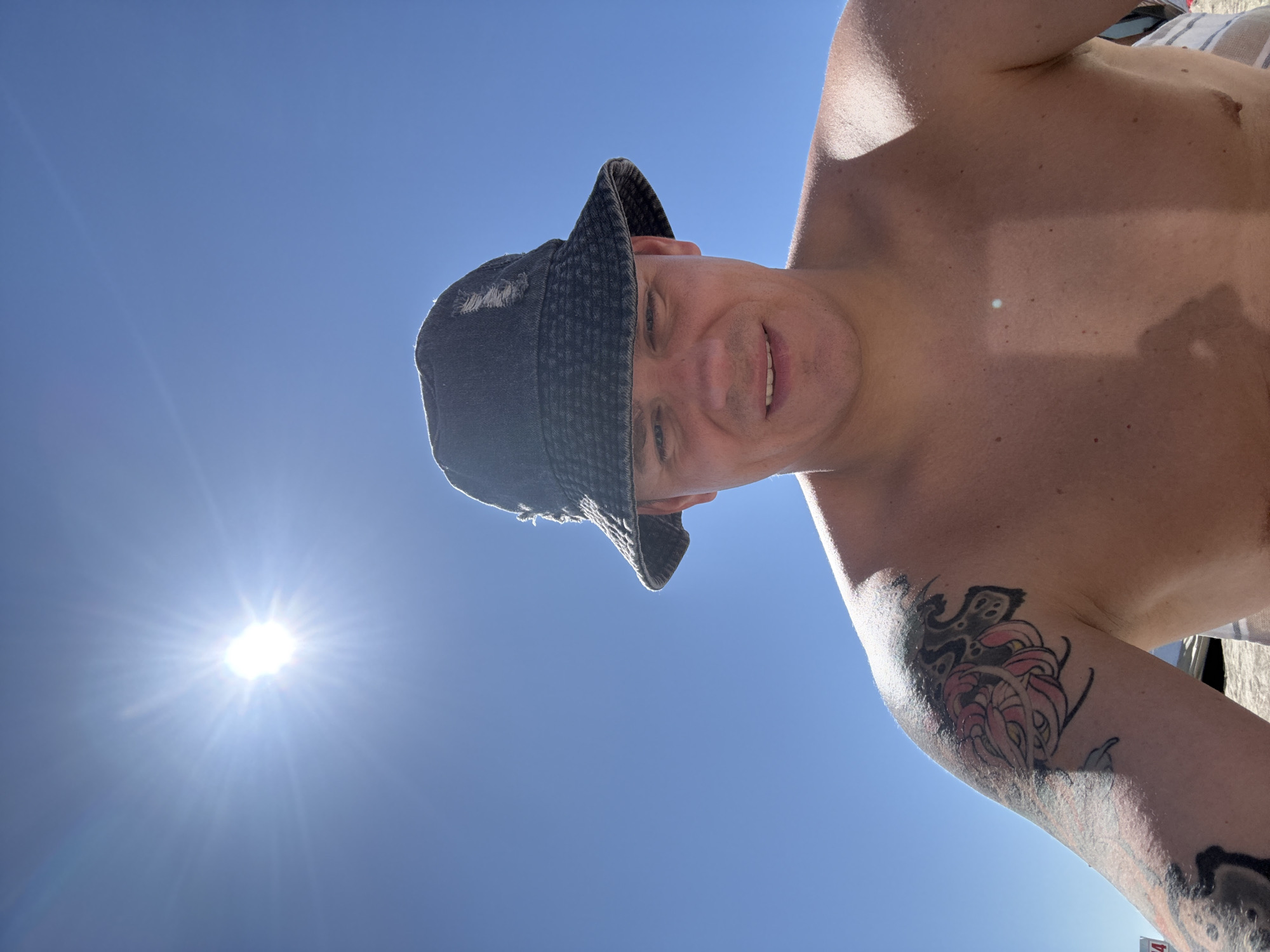This is a close-up, eye-level, outdoor selfie taken on a sunny day. The subject, a bare-chested person wearing a dark bucket hat, is looking directly at the camera. The bright sun is high in the clear blue sky, causing a lens flare and casting strong light. The person has tattoos on their left shoulder and arm. The background appears to be a beach, with sand visible at the bottom of the frame and some blurry elements of what could be beach gear. The specific location of Nakhodka, Russia, cannot be determined from the visual information in the image.