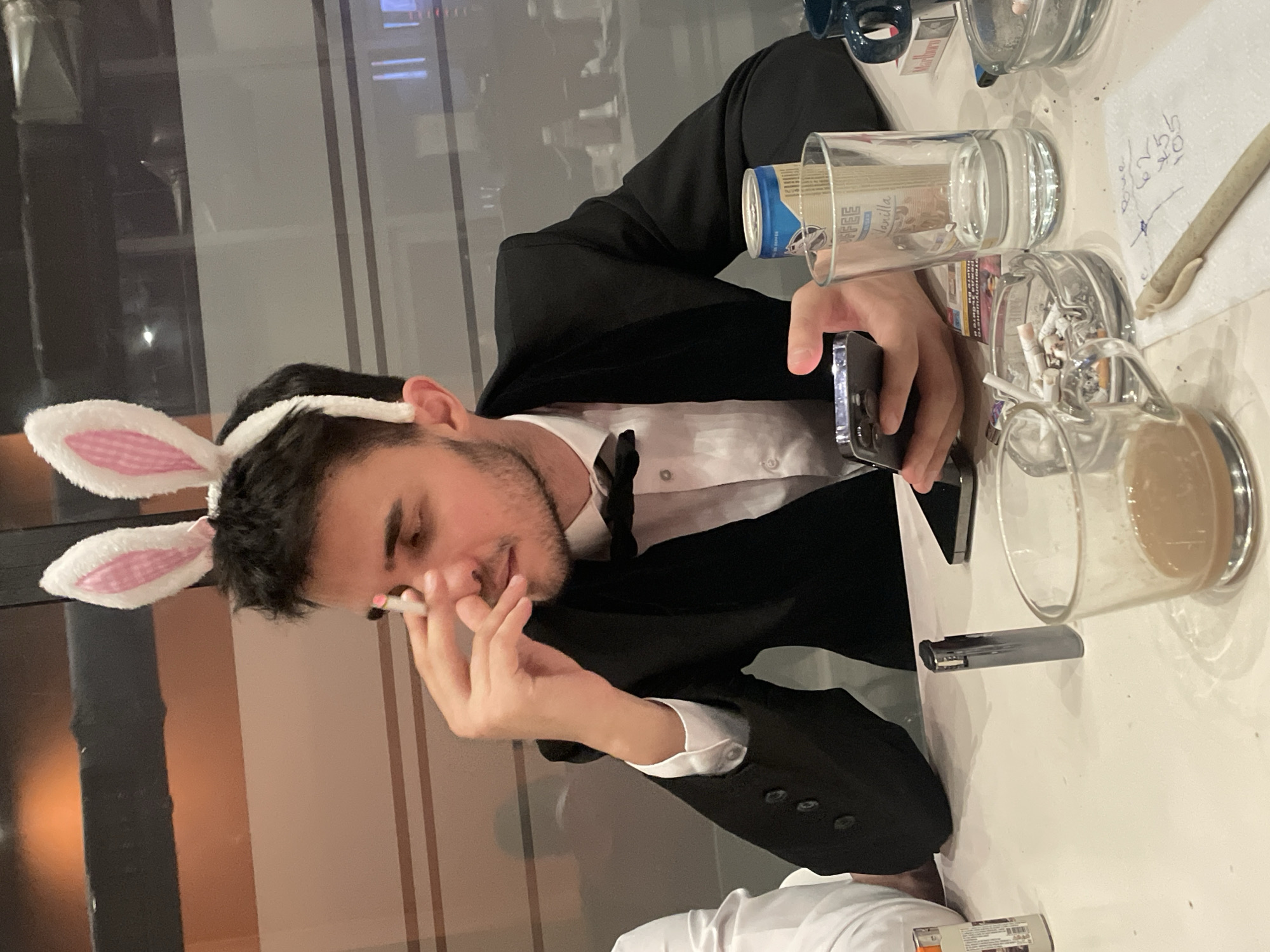 The image, oriented correctly, shows a young man seated at a light-colored table indoors, likely in Sofia, Bulgaria, during the evening or night. He has dark hair, a beard, and is wearing a black suit jacket, white shirt, and a black bow tie, adorned with white and pink bunny ears. His expression is relaxed, with a slight smile, as he looks down at a dark-colored smartphone held horizontally in his left hand. In his right hand, he holds what appears to be a lit cigarette or joint.
The table is cluttered with various items, indicating a social or casual setting. These include a clear glass ashtray filled with cigarette butts, a glass containing a light brown beverage (possibly coffee or tea), and another empty glass with an inverted blue can (marked "ΕΕΕ") inside it. A white paper napkin with handwritten Cyrillic text, including numbers such as "6.55," and a rolled white object resembling a cigarette or joint are also visible. A dark gray lighter and a partially visible "Marlboro" cigarette pack under a blue mug complete the tabletop arrangement.
The background features reflections in what appear to be large windows or glass partitions, suggesting an urban environment or a space with a view. The combination of formal attire with playful bunny ears suggests a festive, costume, or themed event, while the visible smoking materials and beverages indicate a social gathering or a moment of relaxation.