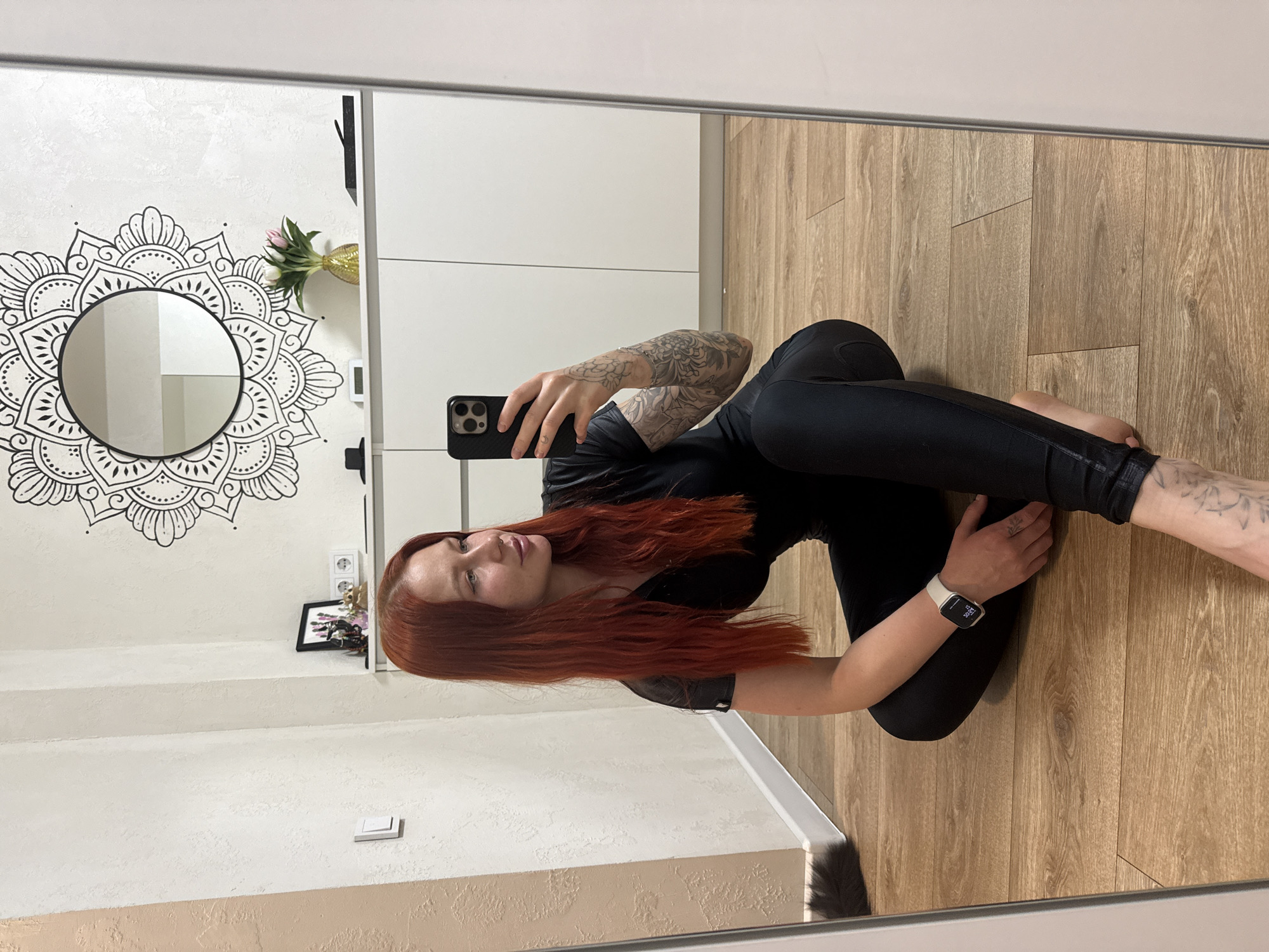 A woman with red hair and multiple tattoos is seen in a mirror selfie inside an apartment in Rostov-on-Don, Russia. She is wearing black athletic leggings and a black top, kneeling on a wooden floor. She is holding a black smartphone in her right hand, taking the selfie, while her left arm rests on the floor. She is wearing a smartwatch on her left wrist.
The background includes a white wall with a large mandala design surrounding a circular mirror. Next to the wall is a white storage unit. A framed picture and a vase with flowers are placed on a surface below the mirror. The mirror's frame is visible. The apartment appears well-lit and modern.