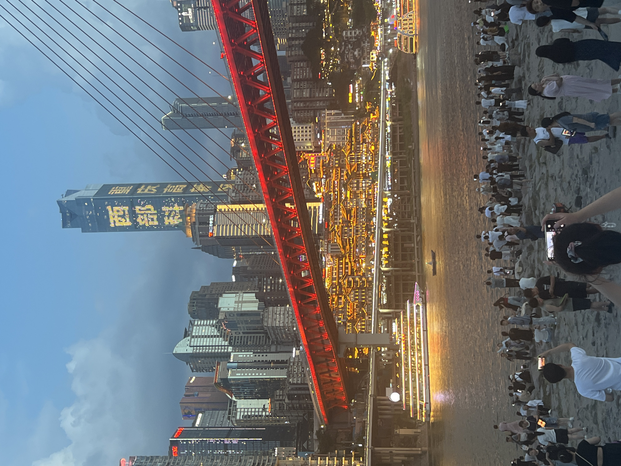 This is a high-angle, full shot of a busy riverside promenade in Yuzhong District, China, likely taken at dusk. In the foreground, a crowd of diverse people are gathered on a rocky shore, some standing, others sitting, and many are holding up their phones, presumably to capture photos or videos of the scene. Several individuals are actively engaged in taking pictures.
Dominating the midground is a large, illuminated red suspension bridge spanning the river. Below the bridge, a multi-story building complex with intricate, traditional Chinese architectural features and glowing yellow lights is visible. A large passenger boat, brightly lit with numerous windows, cruises on the river. Further in the background, the city skyline rises, featuring numerous skyscrapers of varying heights and designs, many adorned with illuminated signage and lights. One particularly tall skyscraper in the center is prominently lit with large Chinese characters. The sky above is a dusky blue with scattered clouds. The overall atmosphere suggests a popular tourist destination with a vibrant evening ambiance.