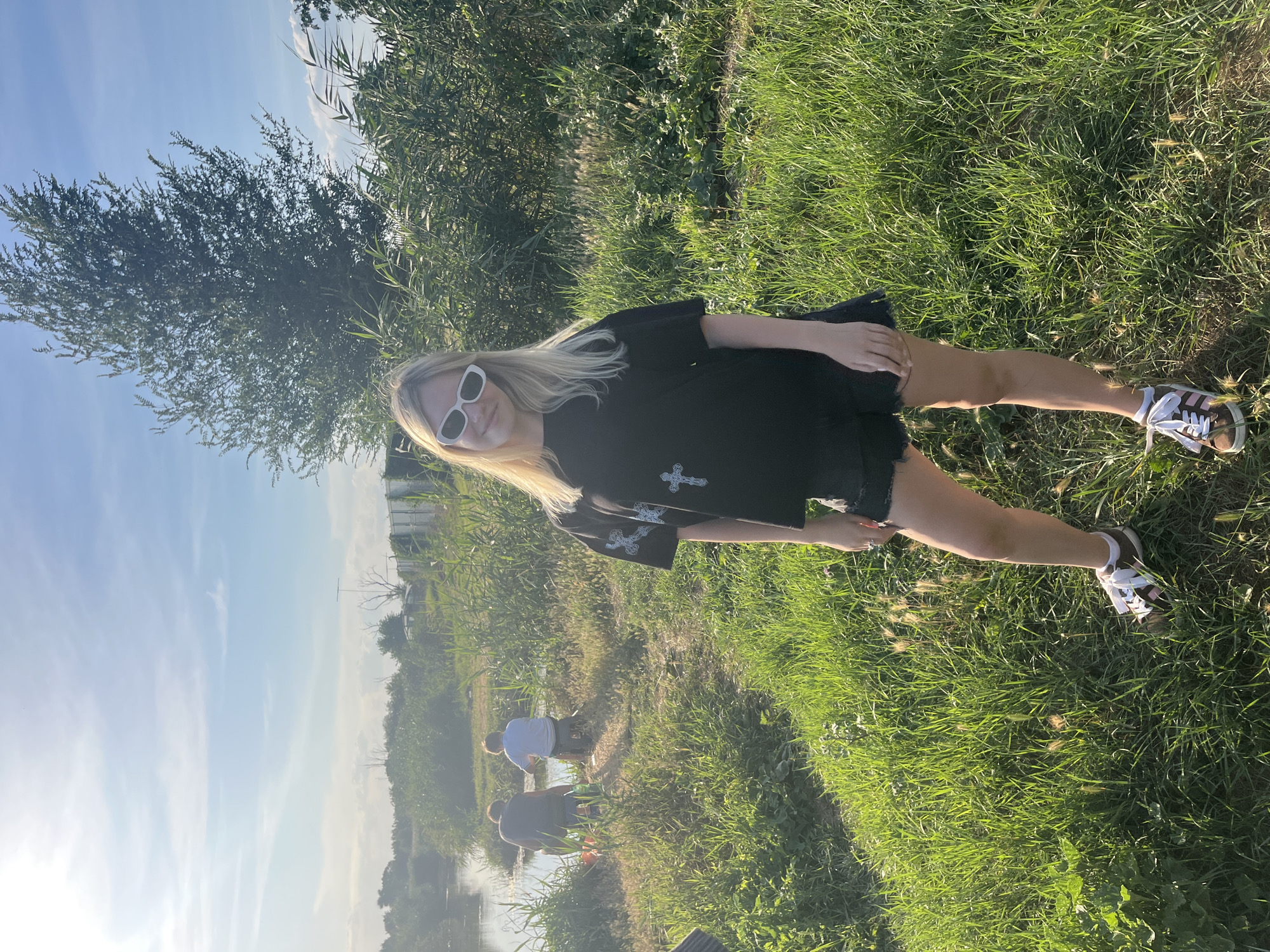 A young woman with blonde hair and white sunglasses stands in a grassy field, looking towards the camera. She is wearing a black t-shirt with a silver cross design and black shorts. She has on white and brown sneakers. In the background, a body of water is visible with two people fishing. The surrounding area is covered in tall green grass and reeds, with a large tree in the distance. The sky is blue with some clouds. The location is identified as Yanovo-Grushevskiy, Russia.