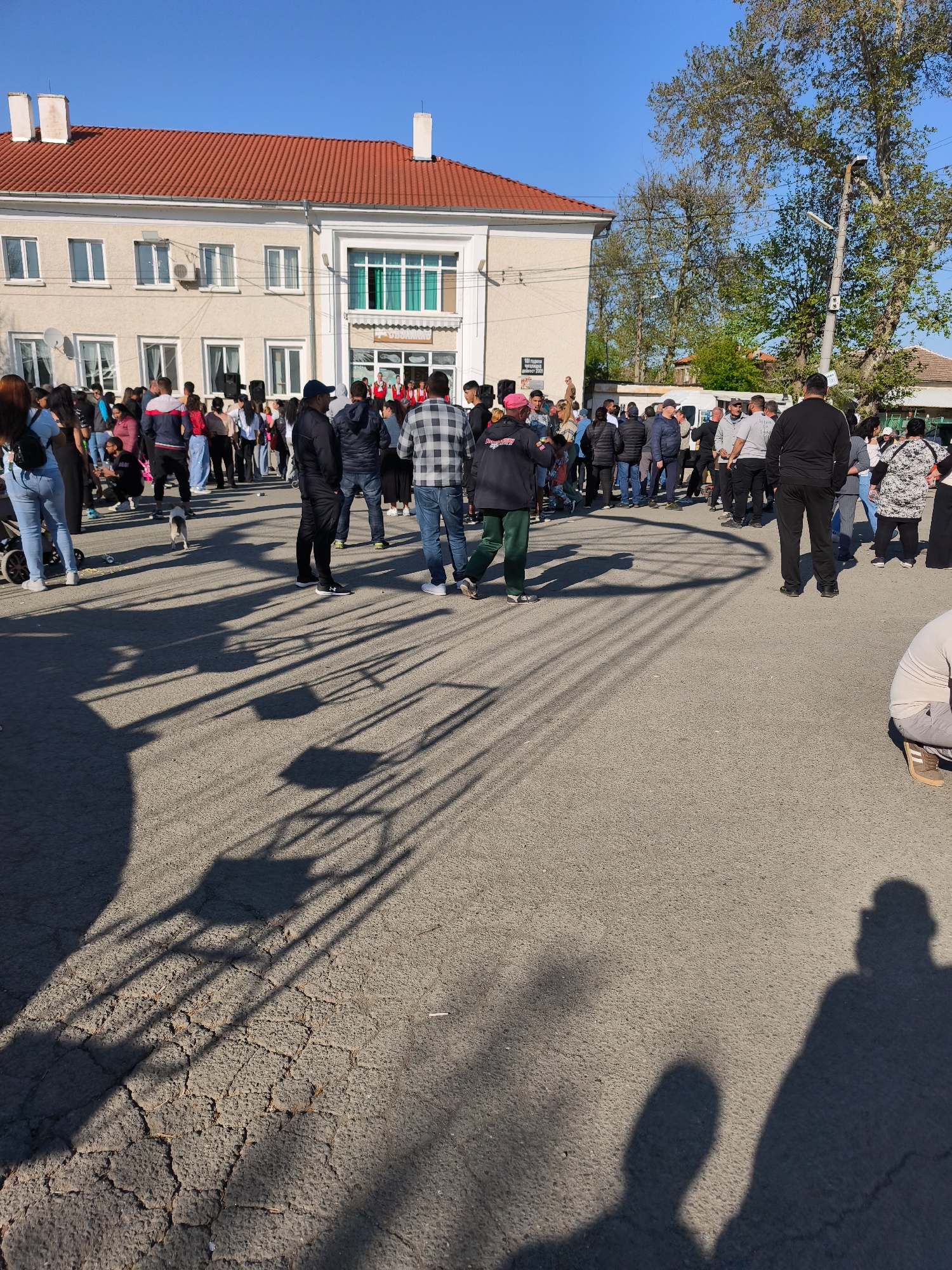 The image depicts a crowd of people gathered in an outdoor area in Rusokastro, Bulgaria. They are standing on a cracked asphalt surface in front of a two-story building with a red tile roof and light-colored exterior walls. Some people are standing in groups, while others are walking. The shadows of trees and poles are cast across the ground. In the background, there are trees and a clear blue sky. The building appears to be a public building. There is a small dog near the center of the crowd. The gathering seems to be centered around a group of people in red and white uniforms, possibly a performance or ceremony.