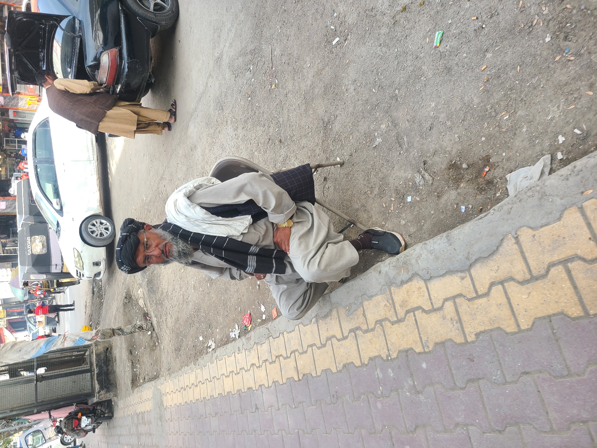The image, taken in what appears to be Quetta City Tehsil, Pakistan, captures a street scene dominated by an older man seated on a simple chair by the roadside. The man has a grey beard, wears glasses, and is dressed in a light grey tunic, a black and white striped scarf around his neck, and a black turban-like head covering. He wears a gold-colored watch on his left wrist and black shoes. He looks directly at the camera with a neutral expression.
In the background, another man wearing a light brown shalwar kameez and a brown waistcoat stands near a black car with its trunk open, seemingly attending to it. A white car is parked nearby. Further in the distance, a black pickup truck with "SUZGAR" written on its side is visible, along with other vehicles and faint figures, including one person in a red shirt.
The ground next to the seated man and the cars is unpaved and dusty, with scattered litter such as plastic wrappers and leaves. A distinct sidewalk with interlocking yellow and purple/brown tiles runs along the bottom and right side of the frame, separating the unpaved area from what appears to be the main walking path. The scene appears to be taking place during the daytime under bright, clear weather conditions.