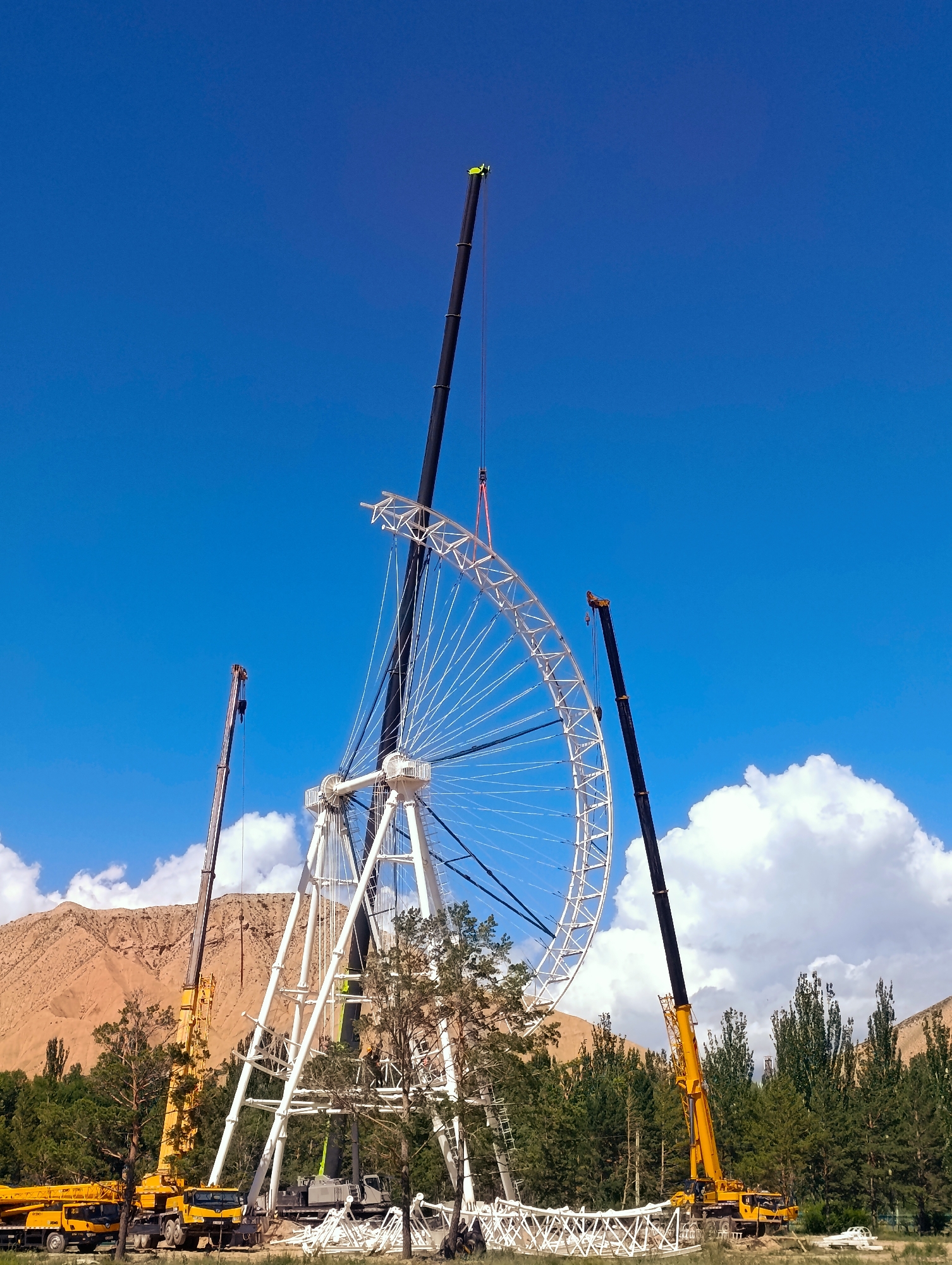 This media file depicts the ongoing construction of a Ferris wheel in Naryn City, Kyrgyzstan. The scene is set against a clear blue sky with scattered white clouds, suggesting fair weather during the daytime. In the foreground, several large yellow cranes are positioned to assist in the assembly of the massive Ferris wheel structure.
The Ferris wheel itself is partially constructed, with a large white wheel framework dominating the center of the image. Multiple white spokes extend from the center, and the outer rim is visibly being pieced together. Two tall, black-armored cranes are actively involved in lifting and positioning sections of the Ferris wheel. A smaller, yellow crane is situated to the left, also seemingly part of the construction effort. In the background, a row of green trees lines a brown, mountainous terrain, characteristic of the Naryn region.
The overall atmosphere suggests a busy construction site with a focus on engineering and infrastructure development. There are no visible people in the frame, but the presence of the heavy machinery clearly indicates human activity. The image captures a moment of progress in building a significant recreational attraction.