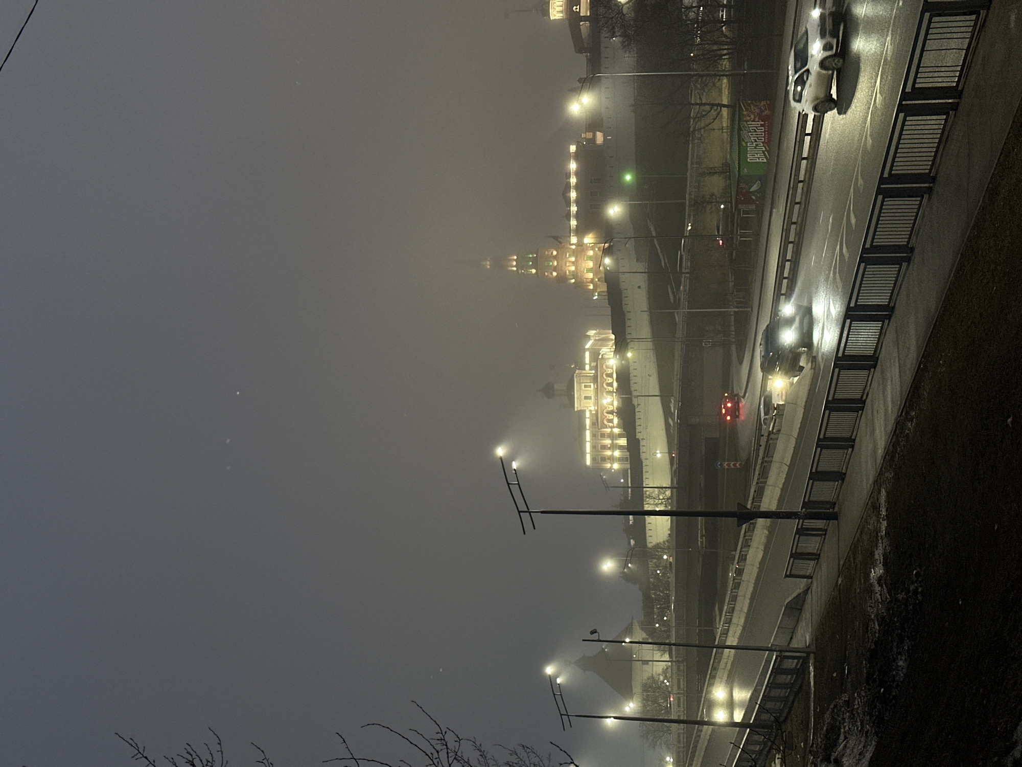 A nighttime scene in Kazan, Russia, captured on a foggy or hazy night. The illumination from streetlights and buildings casts a yellowish glow through the fog. In the background, the illuminated towers and walls of the Kazan Kremlin are visible. A multi-lane road runs along the right side of the frame, with vehicles driving on it. Streetlights line the road, casting light onto the wet pavement. A pedestrian or vehicular barrier separates the road from a grassy area on the far right. The lower part of the frame is slightly obstructed by branches.