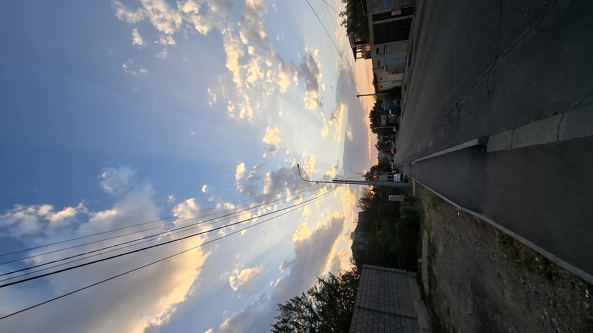 The image captures a street scene in Shymkent, Kazakhstan, at sunset. The sky is a prominent feature, with a vibrant display of blue and scattered clouds illuminated by the setting sun, creating crepuscular rays. Power lines stretch diagonally across the upper portion of the frame. A paved sidewalk runs along the left side of the road, bordered by a low brick wall on the far left and some vegetation. The road itself is asphalt with visible cracks and markings, stretching into the distance. Several cars are present on the road, with one appearing to be stopped at a traffic light. Buildings and structures are visible on both sides of the street, with some appearing residential and others more commercial in nature. The overall impression is of a quiet urban street during the transition from day to night.