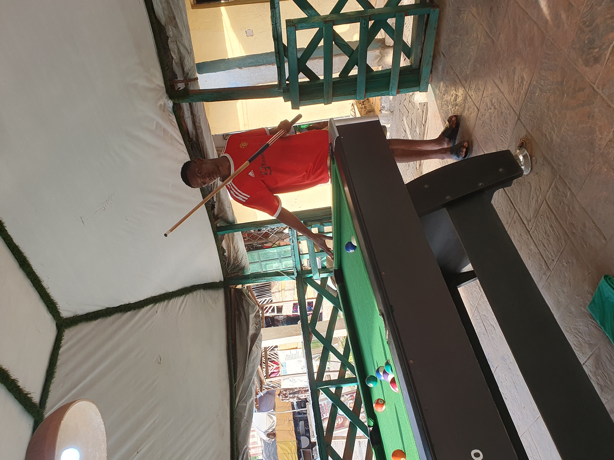 In Lokoja, Nigeria, a young man in a red Manchester United jersey stands beside a pool table, cue stick in hand, ready to play. The pool table is green felt, with several balls scattered across it. The scene is outdoors, under a white tent-like roof, with moss-lined supports. The background shows a marketplace with various stalls and a parked truck. The ground is paved with textured tiles, and a green cloth lies on the floor near the pool table.