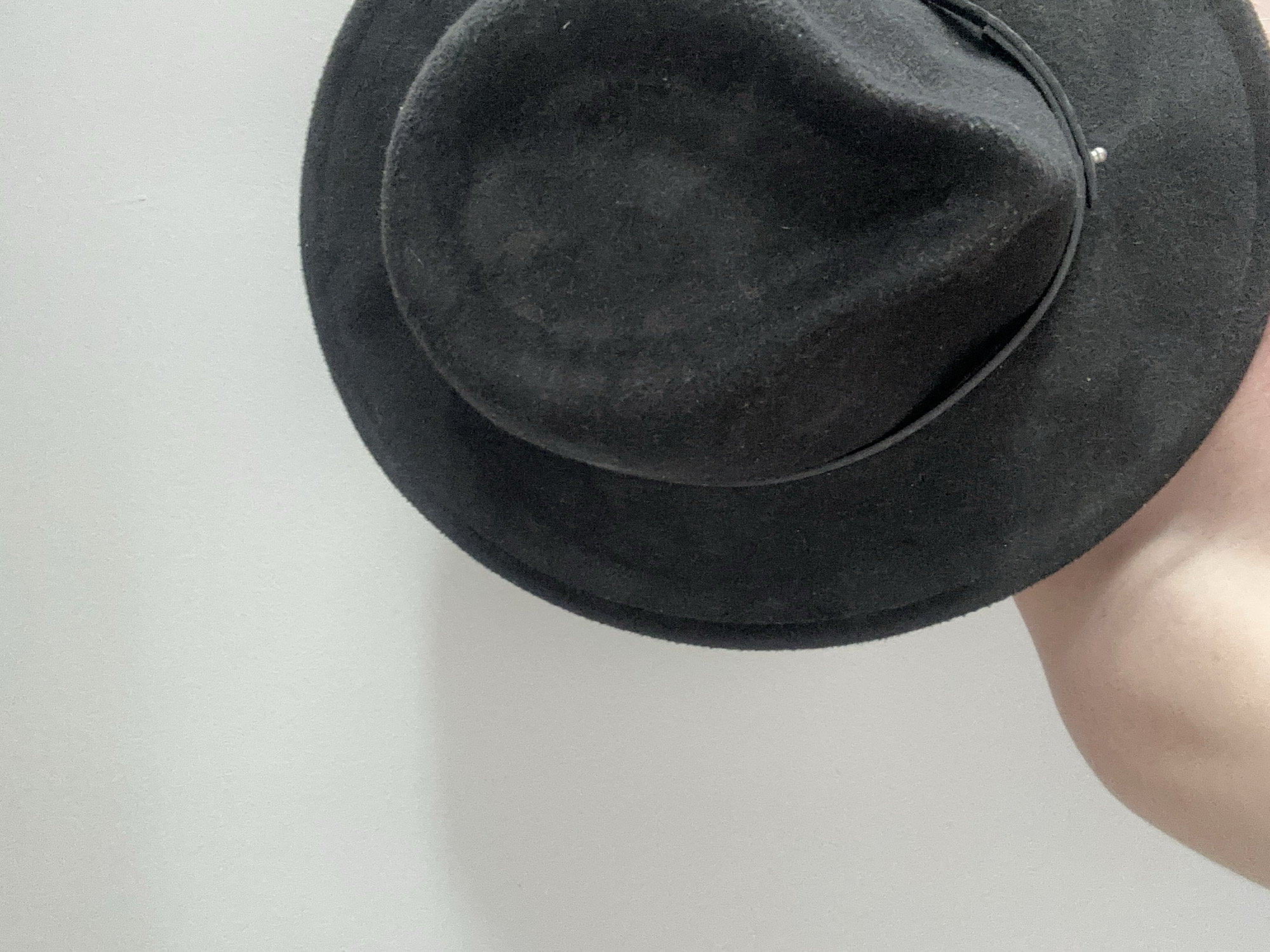 The image is an extreme close-up of a person's shoulder and the brim of a black felt hat. The hat is positioned to obscure the person's face. The background is a plain, light-colored wall. There are no discernible landmarks or elements that would indicate a specific location within Zvezdara Urban Municipality, Serbia.