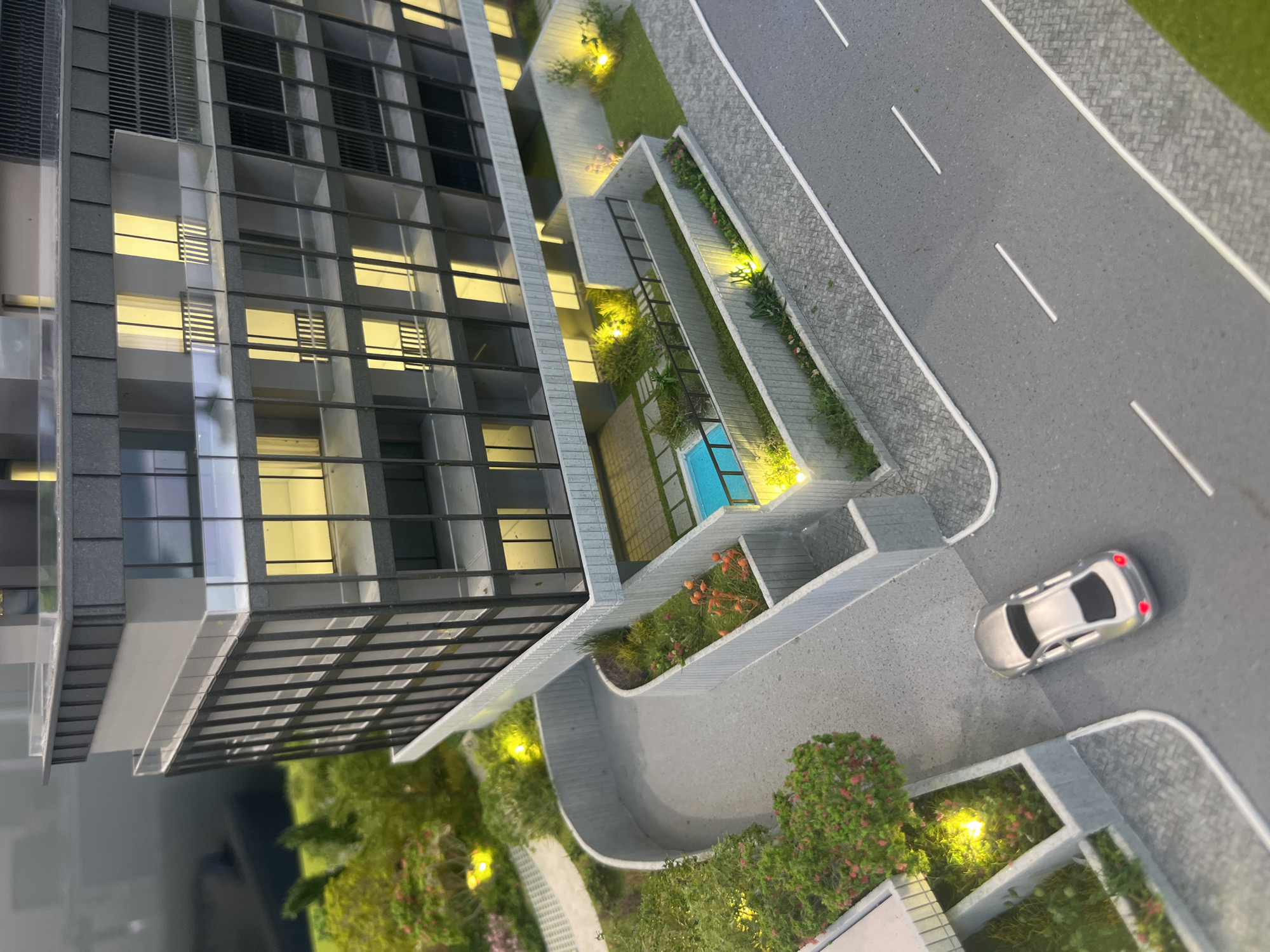 This is a photograph of an architectural model of a modern apartment building in Limassol, Cyprus. The model depicts a multi-story building with dark gray facade elements, large windows, and balconies. In front of the building, there is a paved area with a small swimming pool, surrounded by landscaping with miniature trees and flowers. A silver toy car is parked on a gray road that runs alongside the property. The scene is illuminated by yellow lights within the building and landscape features, suggesting an evening or nighttime setting. The overall impression is one of a high-end residential development.