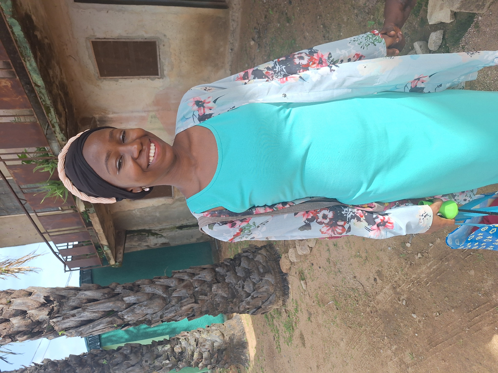 The image shows a woman smiling at the camera, standing outdoors near a building. She is wearing a teal dress and a floral patterned shawl. She has a black headband and appears to be holding a green ball in her right hand. The background includes a weathered building with a small window and a palm tree. The ground is dirt. The location is likely Sabon Tasha, Nigeria, based on user prompt.