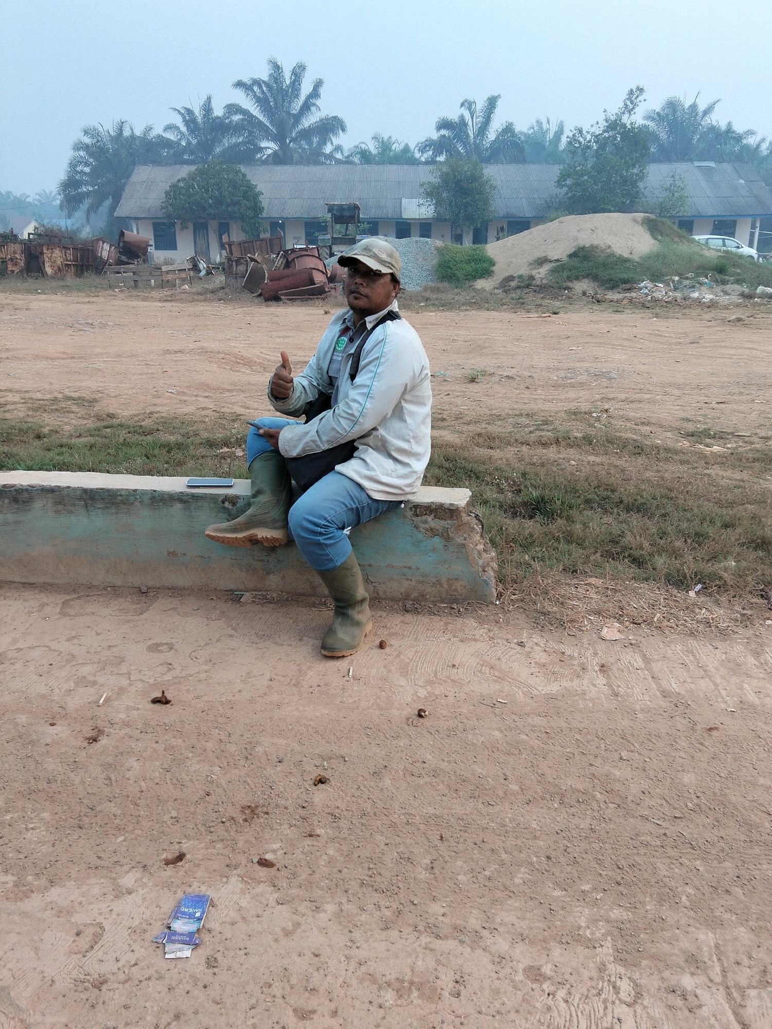 A man wearing a light-colored long-sleeved shirt, blue jeans, a camouflage baseball cap, and tall green rubber boots sits on a concrete ledge. He is giving a thumbs-up gesture and holding a mobile phone in his left hand. The background features a row of single-story buildings with corrugated metal roofs, palm trees, and a pile of sand. The ground is dirt and gravel. The scene appears to be an outdoor industrial or construction area.