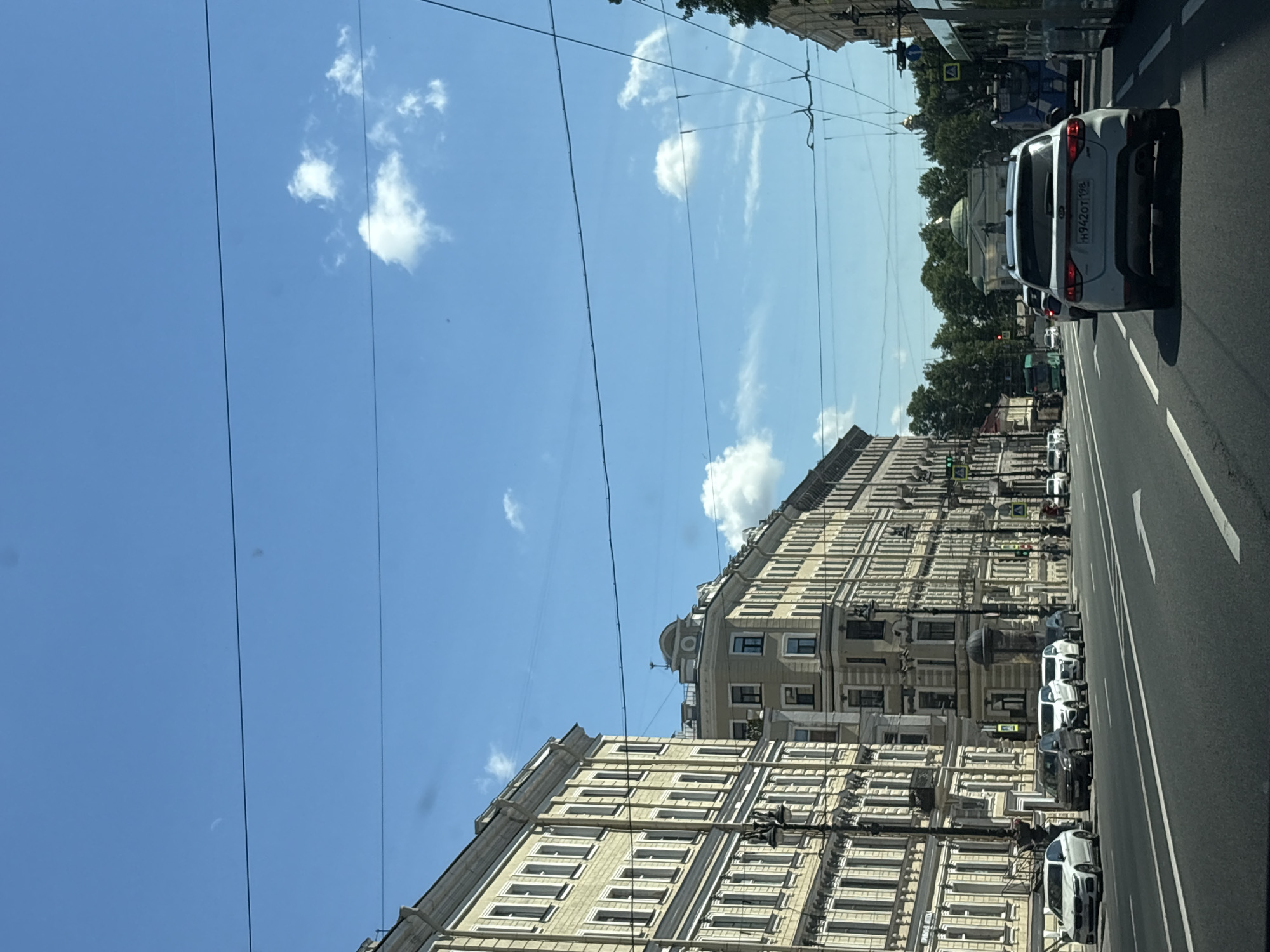 The image captures a street view in Saint Petersburg, Russia, likely on a sunny day with a clear blue sky and scattered clouds. The scene depicts a wide city street with multiple lanes of traffic and parked cars lining the side of the road. Dominating the left side of the frame are ornate, multi-story buildings with classical architectural elements, characterized by numerous windows and decorative facades. Similar architecture continues along the right side of the street, albeit less prominently featured.
Several vehicles are visible on the road, including a white Kia SUV in the foreground, a white sedan, and other cars that appear to be common models. The street itself is paved and marked with lane dividers. Overhead, power lines and possibly tram wires crisscross the sky, a common sight in urban environments. In the background, trees suggest a park or green space beyond the immediate cityscape, and a tram or bus is partially visible on the right. The overall impression is that of a bustling urban thoroughfare in a historic district of Saint Petersburg.