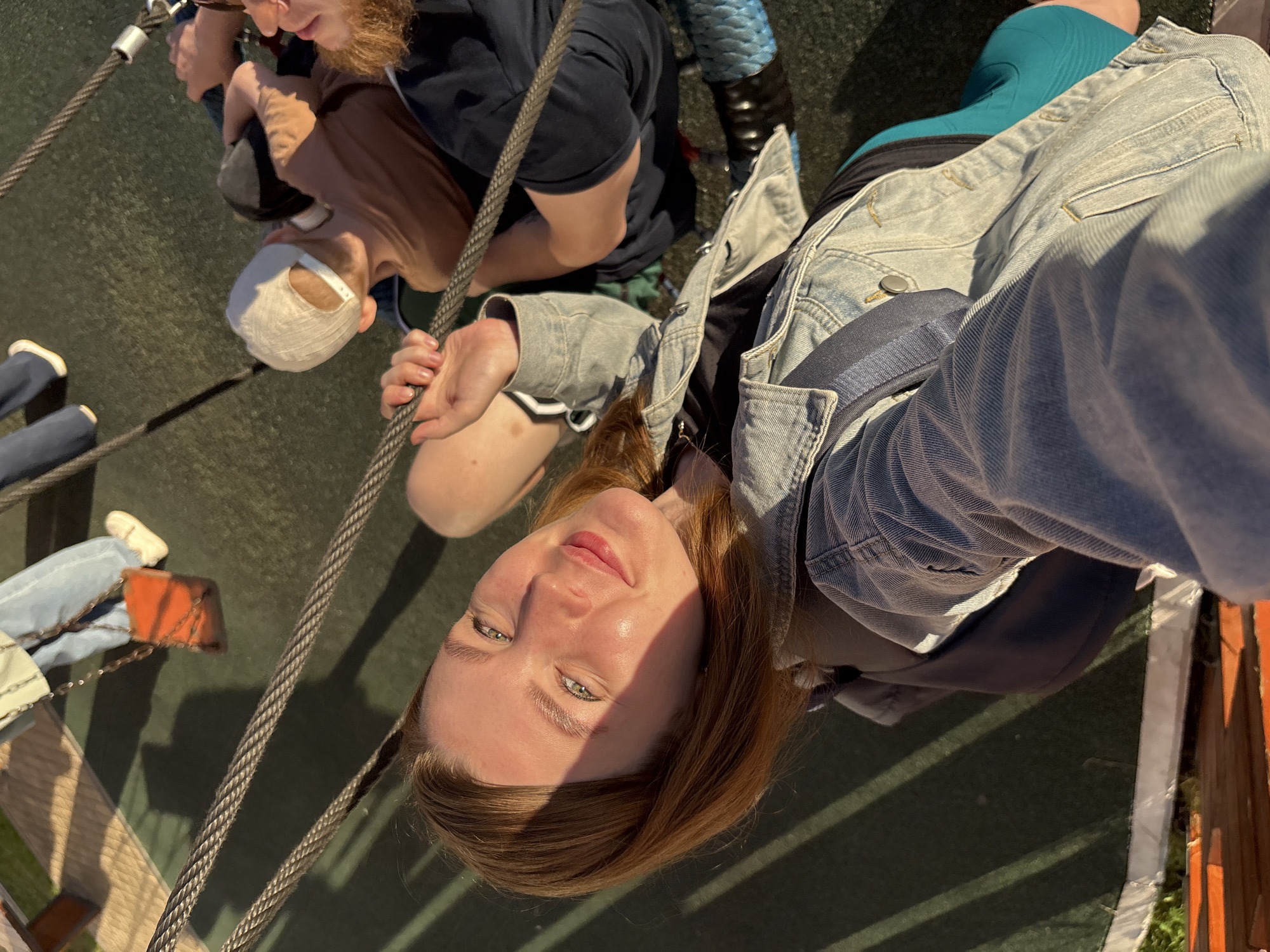 The image is a selfie taken from a high angle, looking down at the subject who is smiling at the camera. The subject is a young woman with shoulder-length auburn hair, wearing a denim jacket over a dark top and turquoise shorts. Sunlight casts a diagonal shadow across her face. In the background, other individuals are visible, including a person wearing a white baseball cap and a tan shirt, and another person with red hair and a beard wearing a dark blue shirt. They appear to be on a playground, with thick metal cables in the foreground suggesting a zipline or suspension bridge structure. The ground beneath them is dark green, possibly rubberized safety surfacing common in playgrounds. A person's legs in light blue jeans and white sneakers are visible in the upper left, near an orange swing seat. The overall environment suggests an outdoor recreational area. The location is identified as a playground in Izhevsk, Russia.