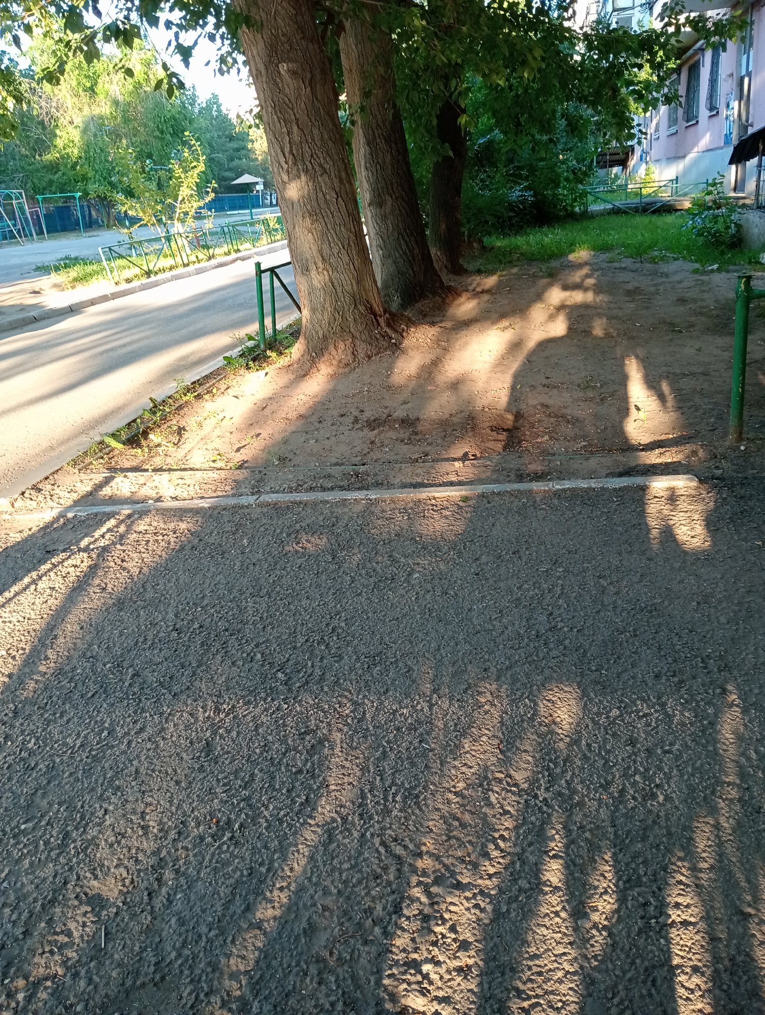 The image shows an outdoor scene with trees and shadows cast on the ground. In the foreground, there is asphalt with dappled sunlight and shadows. A raised curb separates the asphalt from a dirt path where more tree shadows are visible. To the left, a paved road runs alongside a grassy area bordered by a green metal fence. Beyond the fence, playground equipment can be seen. Further in the background, there is a building with a light pink facade and some trees and foliage. The time of day appears to be late afternoon or early evening, judging by the long shadows. The lighting suggests it is a sunny day. The scene looks like a residential area or a park with a playground. There is no visible text in the image.