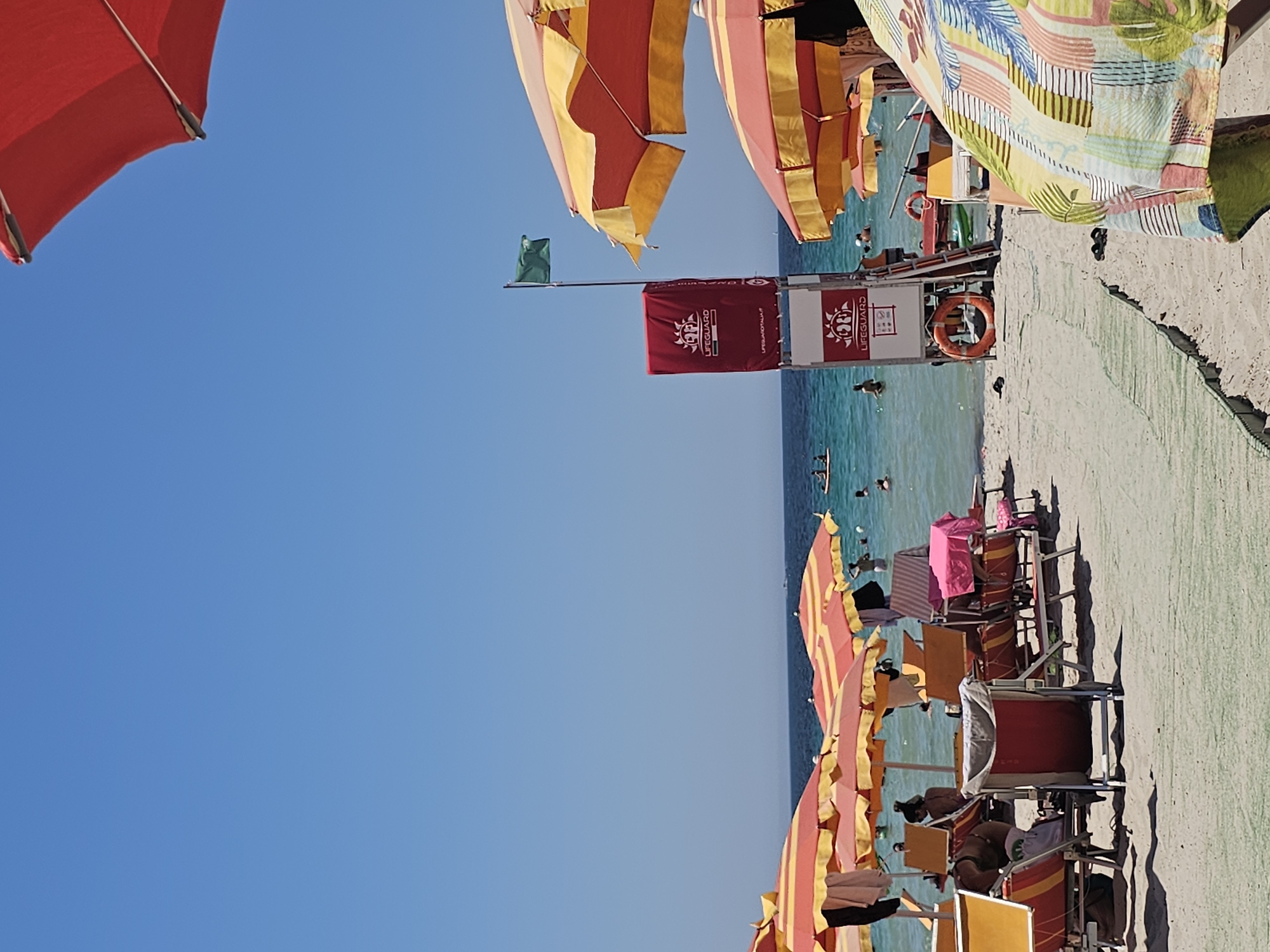 The image shows a sunny beach scene with several people visible. There are many beach umbrellas, mostly red and yellow striped, providing shade. Beach chairs are set up on the sand, some occupied by people. In the background, the turquoise sea is calm with a few individuals swimming or wading.
A lifeguard station is visible with a red flag and a lifebuoy. There are also some towels and bags on the beach. The sky is a clear, bright blue, suggesting a pleasant, sunny day. The overall atmosphere appears relaxed and typical of a summer beach day. The orientation of the image is such that the horizon is vertical on the right side of the frame.
There is some visible text on a red banner that says "LIFEGUARD" and a circular logo with what appears to be a trident. Another white sign with red text and symbols is also visible next to the lifeguard banner. The time of day appears to be daytime, likely mid-morning or afternoon, due to the bright sunlight and shadows. The location appears to be a beach resort or public beach.