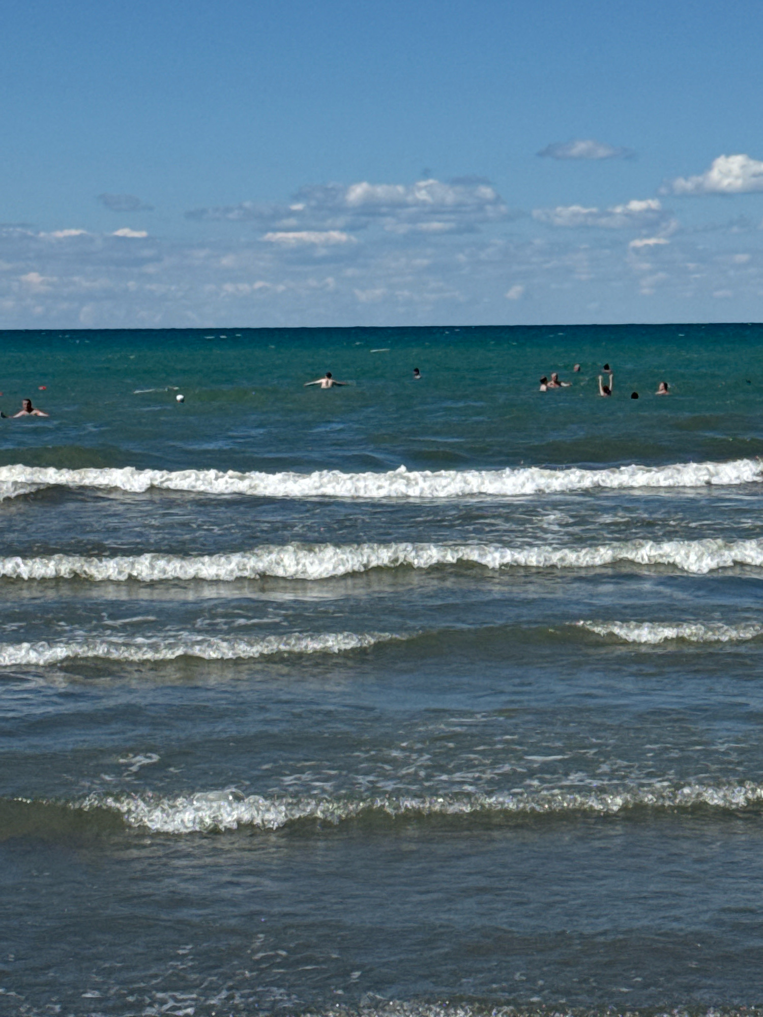 The image shows a view of the Black Sea with several people swimming in the water. The water is a deep turquoise color, and small waves are breaking on the shore. The sky is blue with a few scattered white clouds. The scene is likely Anapa, Russia, a popular resort town on the Black Sea coast, known for its sandy beaches and shallow waters.