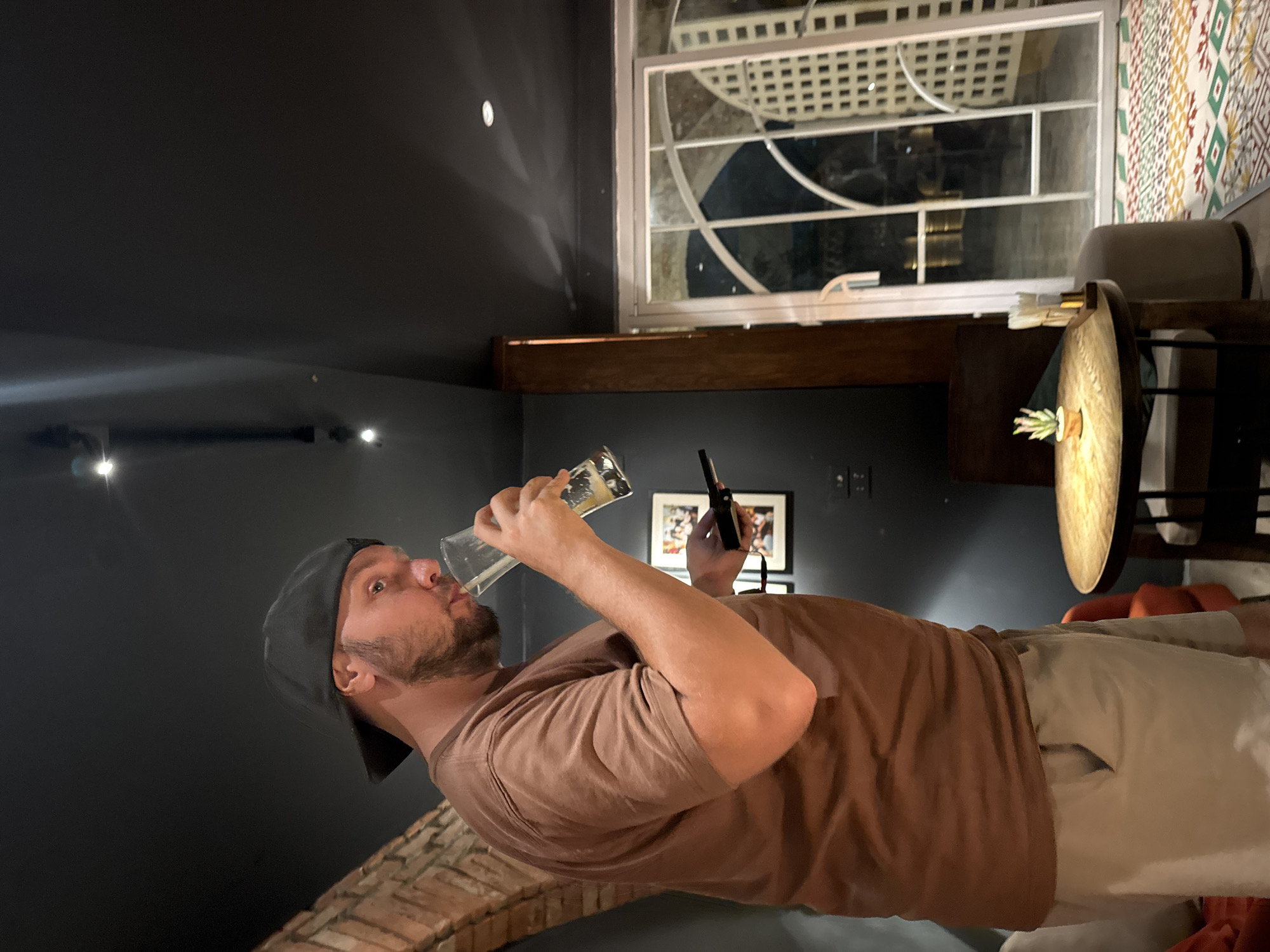The image captures a man wearing a brown t-shirt and khaki shorts, topped with a black baseball cap, as he drinks from a glass of beer. He is standing in what appears to be an indoor setting with dark walls and ceiling, accented by exposed brickwork on the left. In the background, a large window with intricate white latticework allows a view of the night outside. A round wooden table with a small potted plant sits in the foreground, accompanied by chairs and a stool. The man is holding a dark-colored object, possibly a phone, in his left hand. The overall ambiance suggests a casual evening setting, potentially a bar or restaurant in Hà Nội, Vietnam.