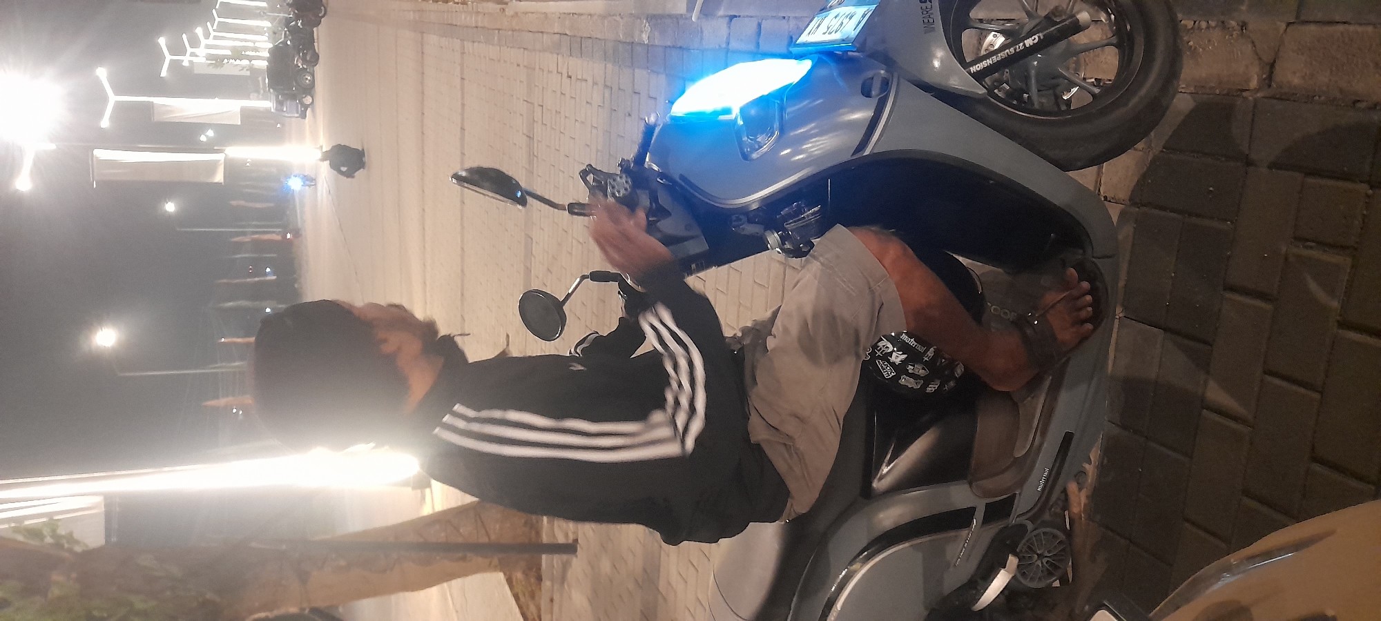 A man wearing a black beanie, a black jacket with white stripes, and light-colored shorts is sitting on a grey scooter at night on a brick-paved sidewalk. The scooter has its headlights on, emitting a bright blue light. In the background, streetlights illuminate a road where other vehicles and a person are visible. A tree is present on the left side of the image. The overall scene appears to be a public area in the city of Kragilan, Indonesia.