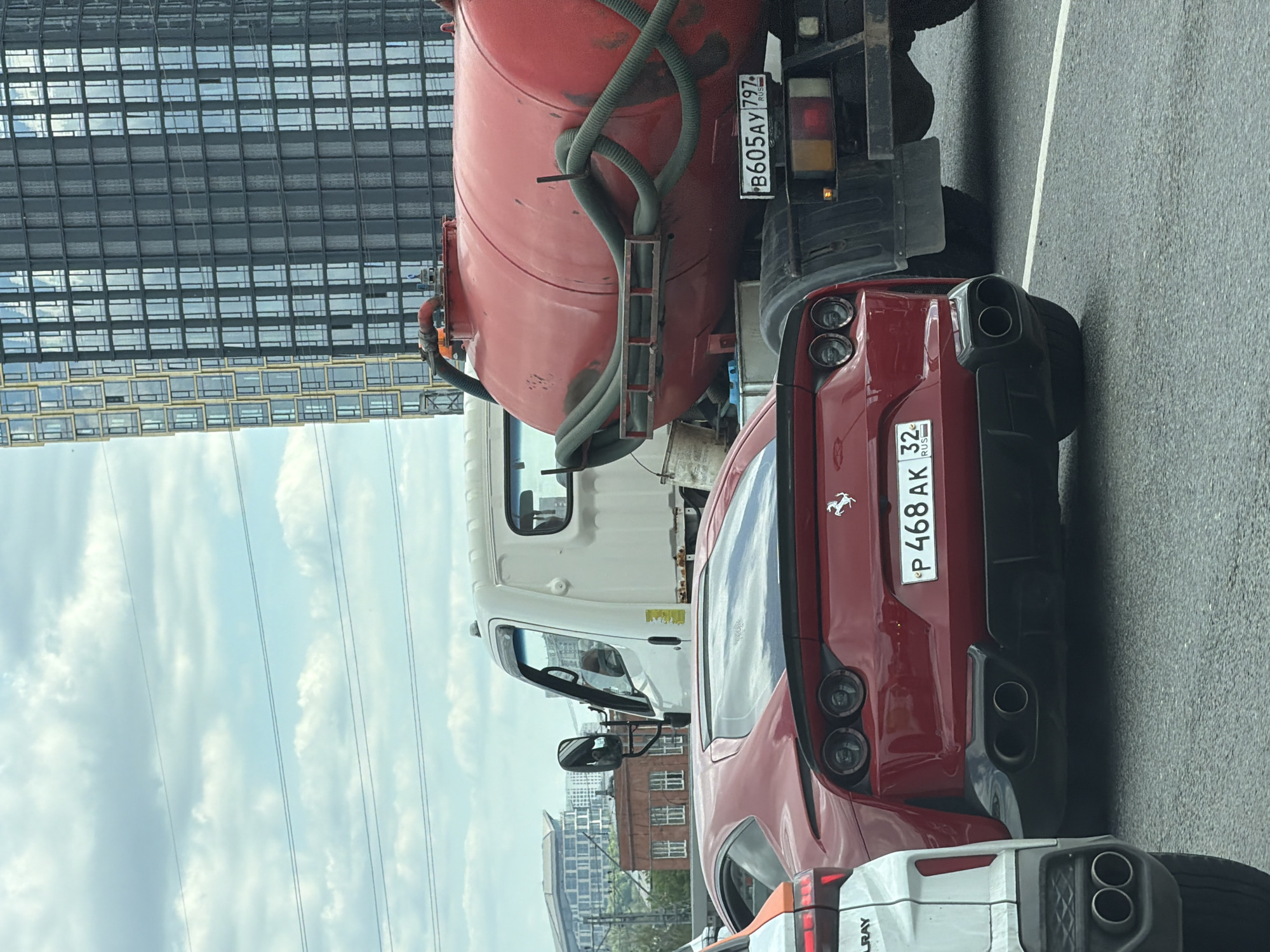 The image captures a moment on a Moscow street. A red Ferrari with license plate P 468 AK 32 RUS is closely positioned behind a white and red truck, license plate B605AY 797 RUS. The Ferrari is followed by a partially visible white vehicle. In the background, modern architecture dominates the skyline. The sky is partially cloudy.