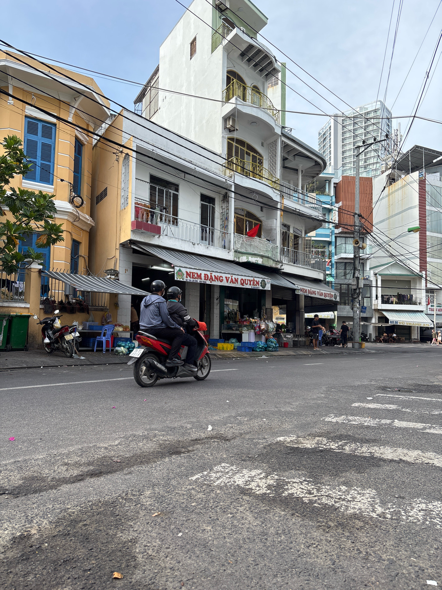 In Nha Trang, Vietnam, a street scene unfolds with a prominent two-story yellow building on the left, featuring blue louvered shutters. Adjacent to it, a multi-story white building showcases curved balconies with gold railings, topped by a white structure with a green trim. Further in the background, modern high-rise buildings rise against a cloudy sky. The street is lined with shops, one with a red flag visible. A red scooter with two passengers wearing helmets travels on the asphalt road, which exhibits wear and tear, including patches and markings. The sidewalk area displays green bins, parked motorcycles, blue crates, and piles of what appear to be watermelons and bags. Overhead, a complex network of electrical wires crisscrosses the scene.
