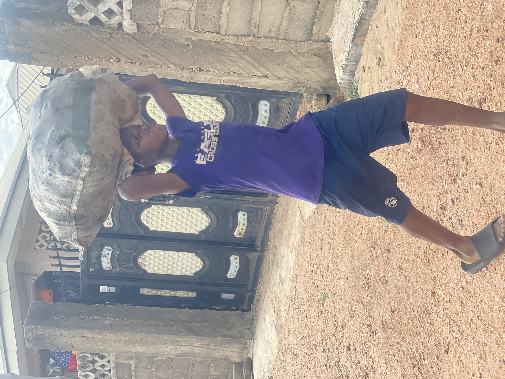 An outdoor shot shows a man in Lokoja, Nigeria, carrying a large, heavy sack on his head. He wears a purple t-shirt and dark blue shorts, and is walking on a reddish dirt ground. A dark metal gate with decorative designs is behind him, next to a weathered concrete building.