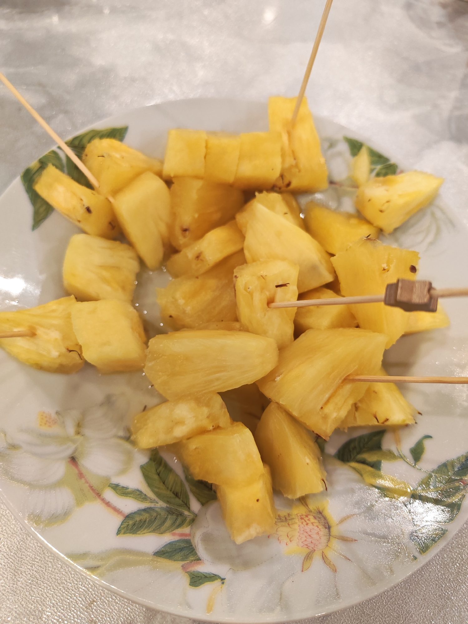 A close-up, overhead shot features a white ceramic plate generously filled with numerous bright yellow, irregularly cut pineapple chunks. The plate is adorned with a delicate floral pattern, showcasing large white flowers with green leaves and subtle yellow or reddish-brown details. Several wooden skewers or toothpicks are inserted into various pineapple pieces, with one skewer featuring a small, square, decorative wooden block. The pineapple pieces appear ripe and juicy, some exhibiting small, dark remnants of the fruit's "eyes." The plate rests on a light-colored, possibly textured tablecloth or shiny surface, which reflects ambient light. The overall presentation suggests the pineapple has been freshly prepared and served, likely as a refreshing snack or dessert, with the skewers facilitating easy individual consumption. No people or visible text are present in the image. The scene appears to be indoors.