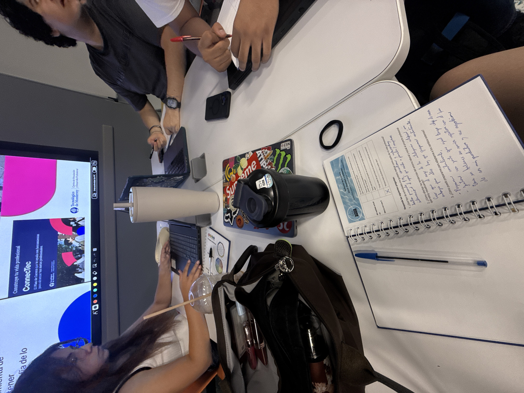 Here is a detailed description of the image:
**Scene:** The image captures a top-down view of a study group at a table.
**People:** Three people are partially visible around the table. One individual is typing on a laptop. One person is seen writing in a notebook.
**Subjects:** The main subjects appear to be the study materials on the table: notebooks, laptops, writing utensils, a water bottle, and a bag.
**Objects:**
- A laptop being used.
- A notebook with handwritten notes, including the word "AGUA" at the top of one page, and questions in Spanish.
- Writing utensils (pens).
- A reusable water bottle.
- A brown bag with personal items inside.
- A cell phone.
- Stickers with the words "SUPREME" on a laptop.
- A screen displaying presentation slides from "Tecnológico de Monterrey," indicating an educational setting.
**Actions:**
- People studying and taking notes.
- Typing on a laptop.
- Drinking from a cup with a straw.
**Interactions:**
- Students collaborating or working individually on assignments.
**Environment:** The scene is indoors in what appears to be a classroom or study area. The lighting is bright. The image is blurry, so it's difficult to discern additional details.
There is no information in the photo indicating it is located in Mexico City, Mexico.