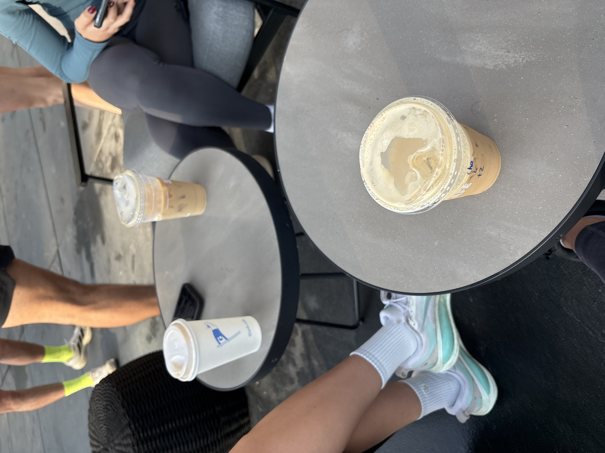 Multiple people are partially visible in an overhead view, primarily showing their legs, feet, and a portion of one person's torso and hands. The scene takes place outdoors, likely at a cafe or patio in Mexico City, Mexico, indicated by the bright daylight and concrete flooring.
Two round, gray tables are central to the composition. On one table, a clear plastic cup with an iced coffee and a lid is prominently displayed, with handwritten text in blue ink that reads "Cha," "<Z," and "2." On the other table, a second clear plastic cup with an iced coffee and a white paper cup are placed. The white paper cup features a blue logo depicting a stylized figure in an inverted pose, with faint text below it, possibly "@radioc_m."
The activity involves people sitting around the tables, drinking coffee, and potentially socializing. One person is seen wearing dark athletic leggings and a teal long-sleeved top, holding a dark smartphone. Another individual wears light-colored sneakers with mint green accents and white ribbed socks. Bare legs and feet with sneakers and bright neon yellow socks are visible from a third person. The overall atmosphere appears casual and relaxed on a bright, sunny day.