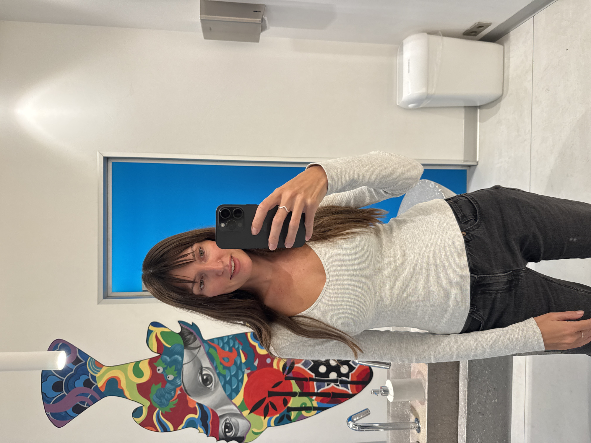 A woman with long brown hair is taking a selfie in a brightly lit, modern bathroom. She is wearing a light gray long-sleeved shirt and dark jeans. The bathroom features a vibrant mural on the wall behind her, depicting abstract shapes and human eyes. To her right, there is a blue door, a silver paper towel dispenser, and a white trash can. A gray sink with a chrome faucet is visible on the left side of the frame. The floor is tiled in a light gray pattern. The overall impression is one of contemporary design and cleanliness. There is no indication that this scene is located in Moscow, Russia.