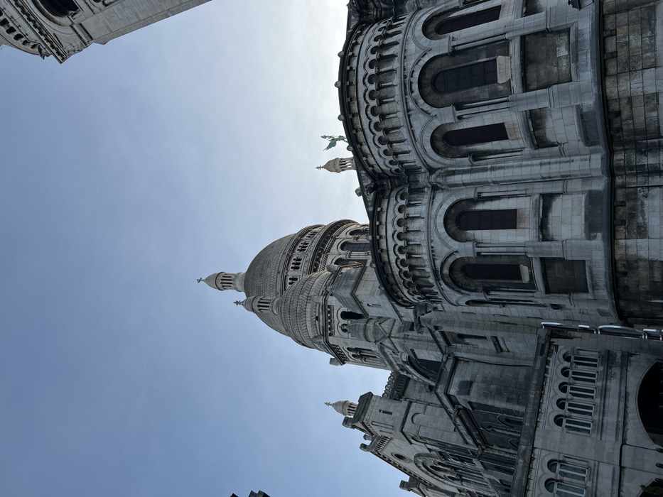 The image displays the Sacré-Cœur Basilica, situated on Montmartre in Paris, France. The perspective is a low-angle upward shot, capturing a significant portion of the basilica's exterior under a clear, light blue sky.
The prominent central dome, topped with a cupola and a cross, dominates the upper-mid section of the structure. To its left and right, smaller domes and turrets are visible, demonstrating the building's Romanesque-Byzantine architectural style. The basilica's facade is constructed from a light-colored, likely travertine, stone, exhibiting intricate details such as arched windows, rounded buttresses, and decorative cornices. A statue, possibly of a figure holding a flag or staff, with a green patina, is visible atop a lower turret on the right side of the main dome. A tall, multi-tiered bell tower is partially visible along the extreme right edge of the frame. The lighting indicates daylight, with soft shadows defining the architectural relief. No individuals or other distinct objects are discernible in the immediate foreground or background, beyond the structure itself.