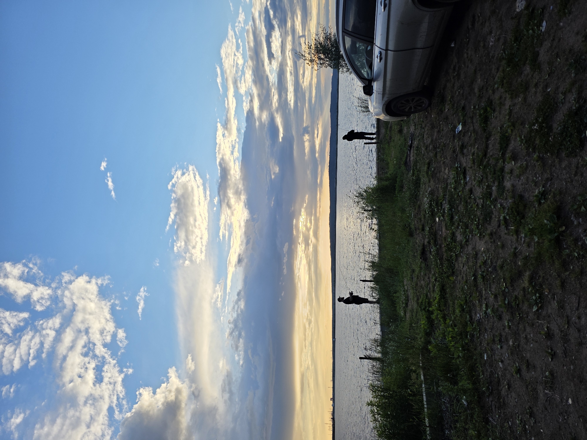 The image depicts a serene waterside scene at dusk in Chaikovsky, Russia. Two figures, silhouetted against the shimmering water, are visible. One stands closer to the shore, appearing to be fishing or engaged in a similar activity, while the other is slightly further out. A silver car is parked on the grassy bank, partially visible on the right side of the frame. The sky is a canvas of blues and yellows, with dramatic, wispy clouds catching the last rays of the setting sun. The water reflects the colorful sky, creating a tranquil and picturesque atmosphere. The ground is a mix of dirt and green grass, with some vegetation growing along the water's edge. The overall mood is peaceful and contemplative, suggesting a moment of quiet enjoyment by the water as day transitions into night. There is no visible text in the image.