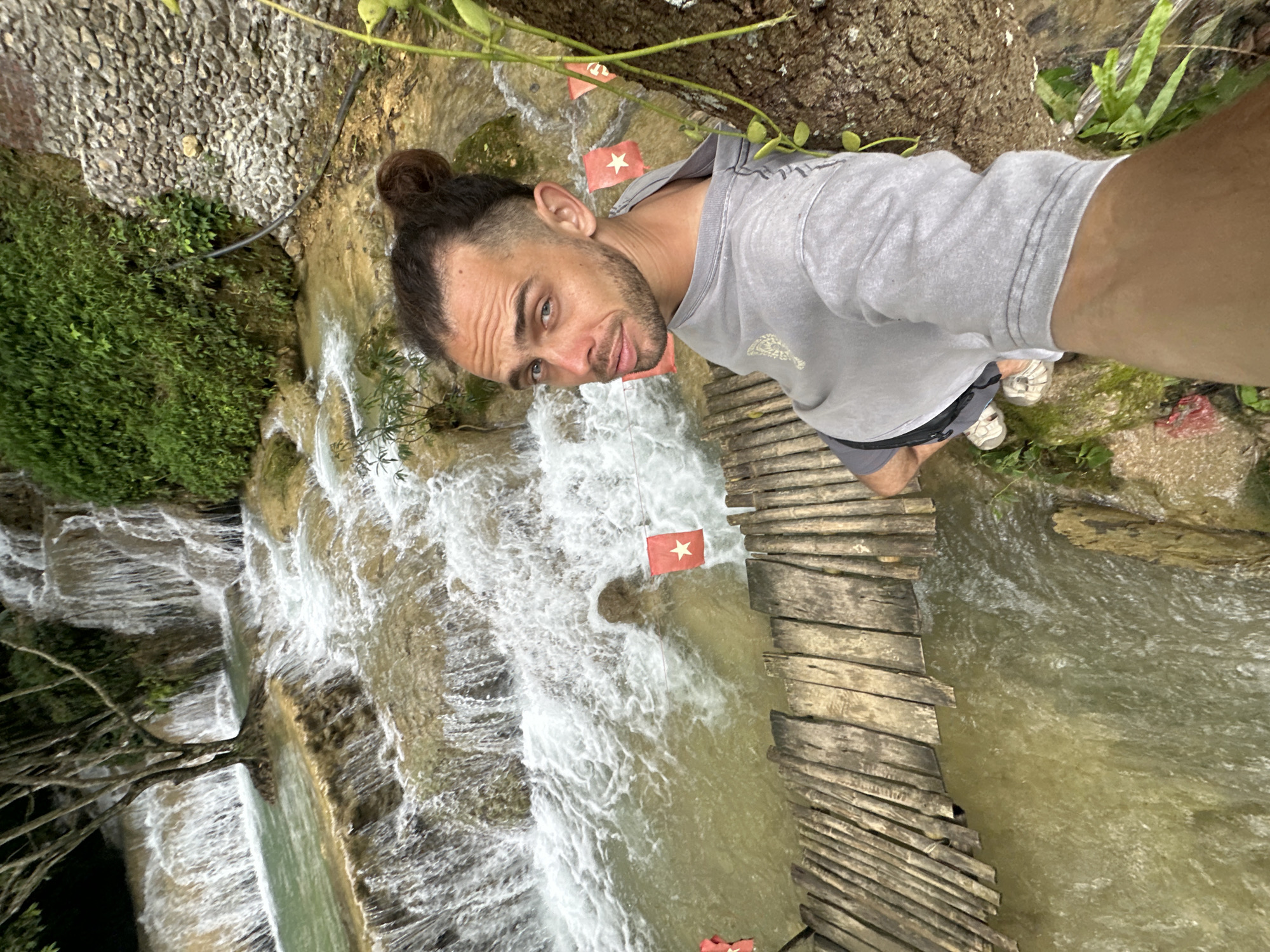 A man with dark hair styled in a bun, and a light beard, is standing on a wooden plank bridge near a multi-tiered waterfall. He is wearing a grey hooded sweatshirt and black shorts. His eyes are looking directly at the camera. The waterfall is surrounded by lush green vegetation and rocky terrain. There are several small red flags with white stars visible, attached to strings that span across the water in front of the waterfall. The water is a murky green color. The bridge is made of rough, weathered wooden planks.