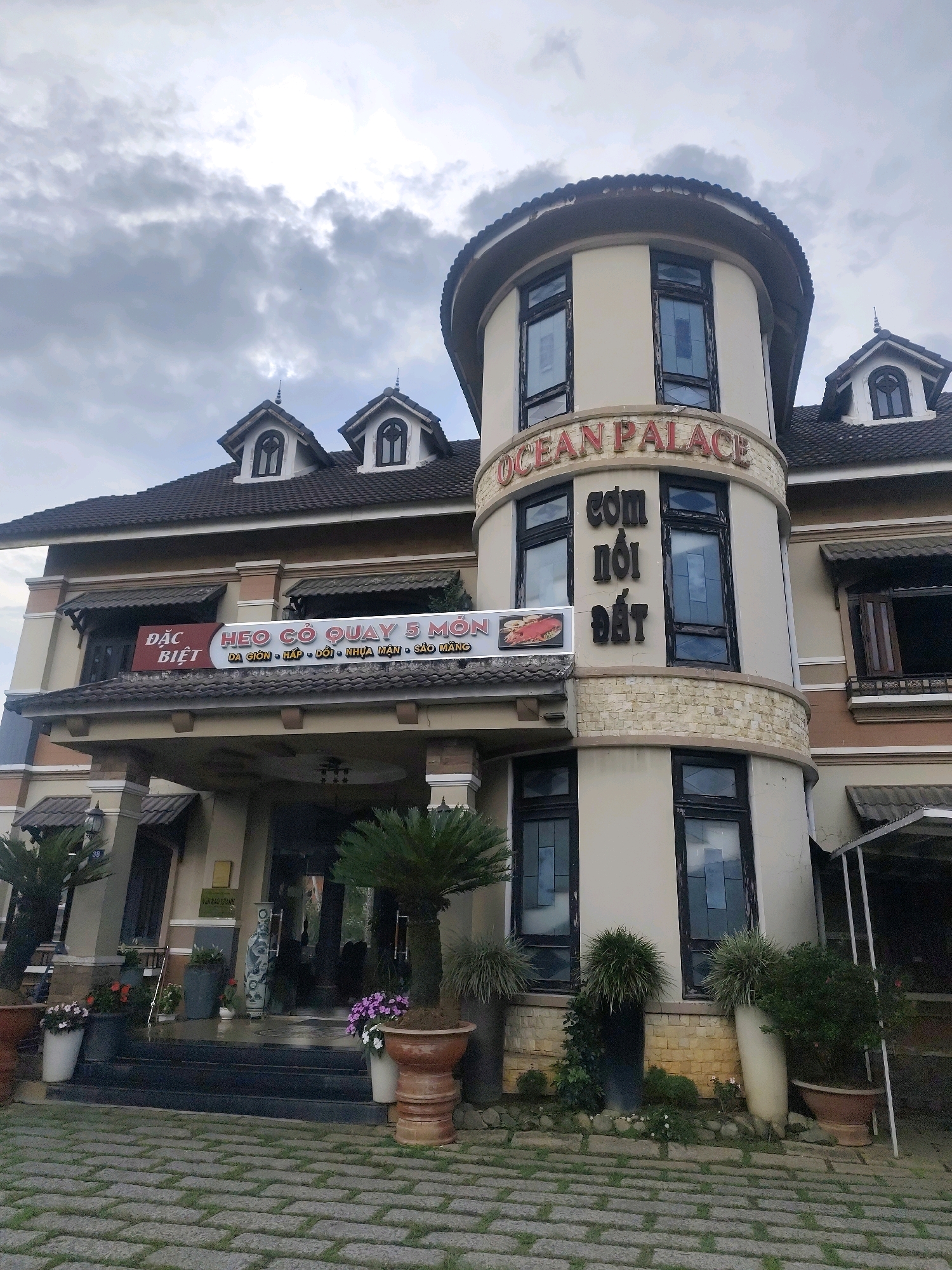 The image shows the exterior of a building identified by the signage "OCEAN PALACE" in red lettering on a rounded tower section. Beneath "OCEAN PALACE" are Vietnamese characters that translate to "COM NOI DAT," likely indicating the establishment is a restaurant. Another sign displays "ĐẶC BIỆT" (Special) and "HEO CƠ QUAY 5 MÓN," suggesting a special dish of roast suckling pig with five preparations. The building features a dark tiled roof with dormer windows and a beige facade. The foreground is a stone pathway leading to a set of stairs and the entrance of the building. Potted plants, including palm-like trees and flowering bushes, are placed around the entrance and the base of the tower. The sky is overcast with dark clouds. The architecture and the presence of a restaurant specializing in local cuisine are characteristic of tourist establishments in Da Lat, Vietnam.