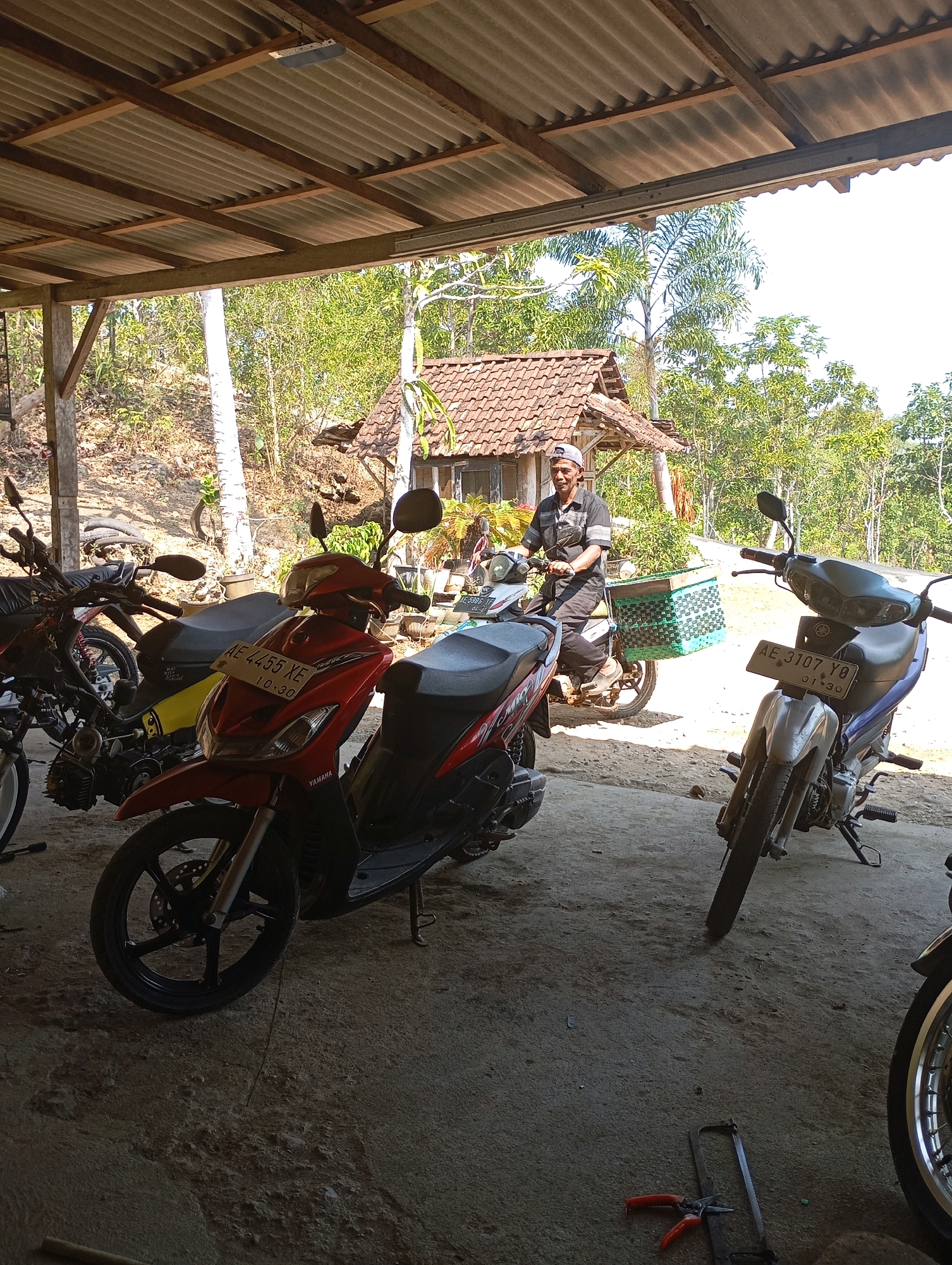 The image captures an outdoor scene under a corrugated metal roof, likely a workshop or garage, in Ketepung, Indonesia. In the foreground, a red scooter is parked, its license plate partially visible. To its right, a silver and blue motorcycle is also parked, its license plate clearly showing "AE 3107 YO". In the background, a man wearing a cap and a dark shirt is seated on another scooter, which is equipped with a green basket. He appears to be looking towards the left. Behind the man, a simple building with a tiled roof is visible, surrounded by lush green trees and vegetation, suggesting a rural or semi-rural setting. The ground is paved with concrete or dirt. Tools, including a saw and pliers, are scattered on the ground in the lower right portion of the image. The overall impression is of a functional outdoor space where motorcycles are stored and possibly maintained.