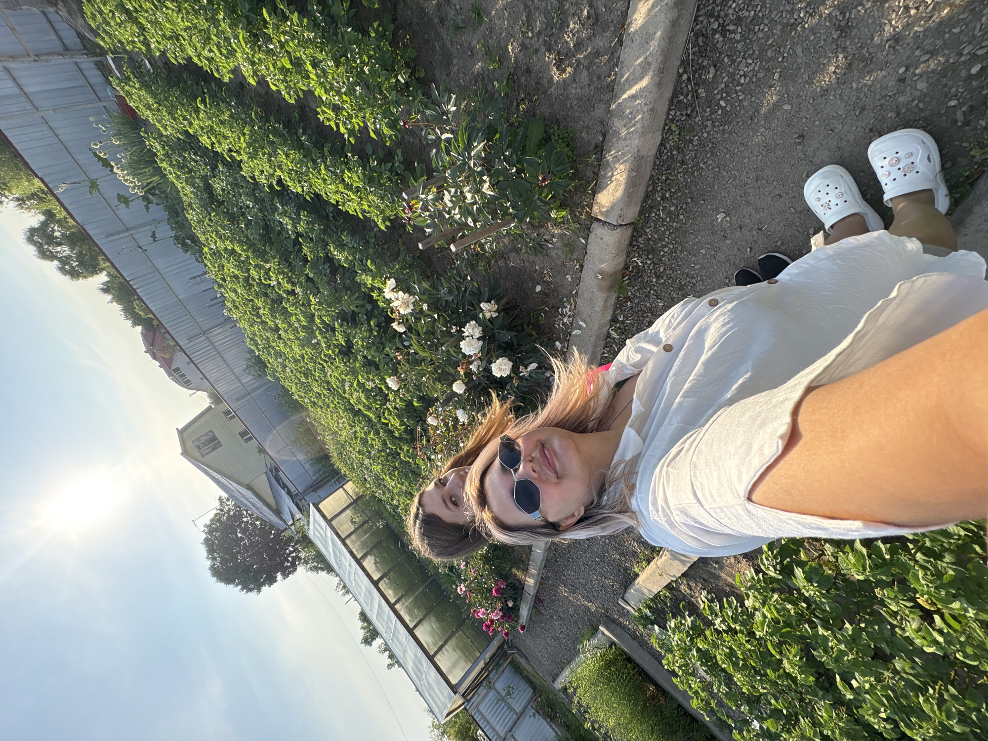 In a garden setting located in Razdolnoye, Russia, two individuals are present. In the foreground, a woman with blonde hair, wearing sunglasses and a white dress, is taking a selfie. She has pink highlights in her hair and is wearing white clogs adorned with flower-like embellishments. To her left and slightly behind her, a young woman with brown hair is looking towards the camera. The environment features rows of green plants, possibly potato plants, stretching up a slope towards a metal fence. A greenhouse structure with a transparent roof is visible to the left. In the background, a house with a light-colored facade and a slanted roof can be seen, along with trees and a clear blue sky illuminated by bright sunlight. The ground is covered in gravel and soil, with some wooden supports and stone edging bordering the garden beds. Pink and white flowers are blooming in some areas of the garden.