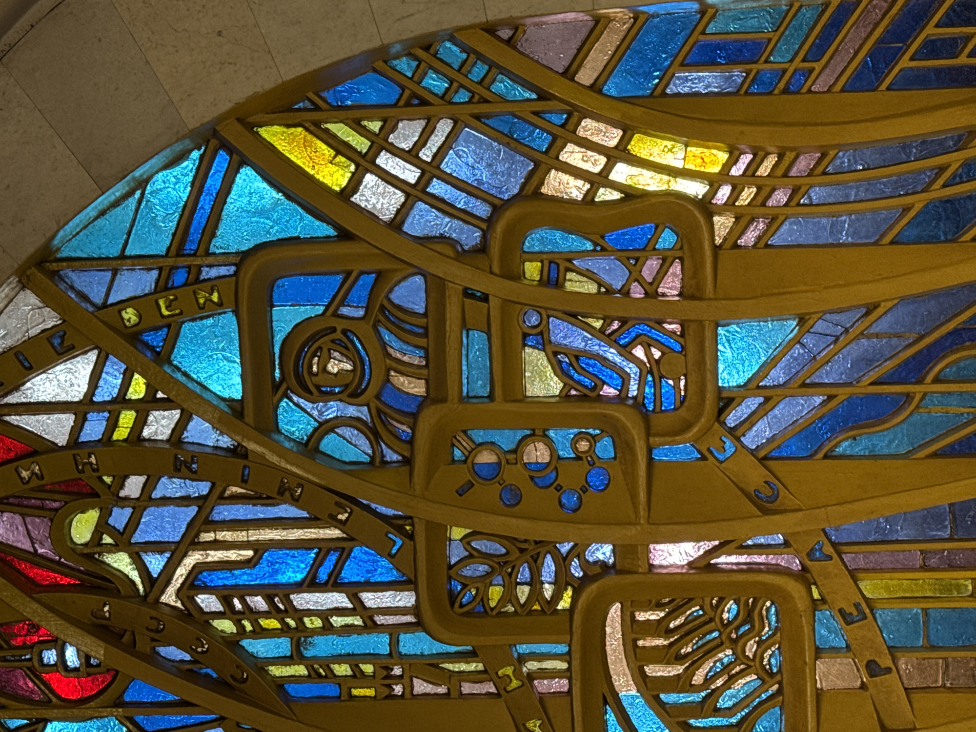 This media file captures a close-up of a vibrant, intricate artwork resembling a stained glass window or a decorative wall panel. The artwork features numerous multi-colored glass pieces, predominantly in shades of blue, golden yellow, and white, with accents of red, purple, and green. These textured glass segments are held together by thick, golden-brown metallic or metallic-looking frames.
The scene is a static architectural detail, likely indoors, given the soft, diffused light passing through the colored glass and the curved, light-colored wall visible in the upper left corner. No activity or event is taking place.
Notable details include a complex design that combines abstract geometric patterns with specific symbolic elements. Several letters are embedded within the golden framework. Parts of the word "SCIENCE" are clearly visible, specifically "S C I E N" and "C E". Another section contains "M I N" and "I N G", suggesting the word "MINING". Other isolated letters like "U", "J", and "Y" can also be identified.
Figurative symbols are also present: one prominent square frame encloses a motif of magnifying glasses and interconnected circular shapes representing molecular structures, strongly suggesting a theme of scientific research or chemistry. Below this, another frame depicts a stylized leaf or botanical pattern. Other areas seem to incorporate mechanical or industrial shapes, such as a potential gear-like element. The overall composition is dynamic and layered, hinting at themes of knowledge, industry, nature, and urban development. The artwork is rich in detail and color, with light enhancing the textures of the glass. The location context indicates this piece is in Moscow, Russia.