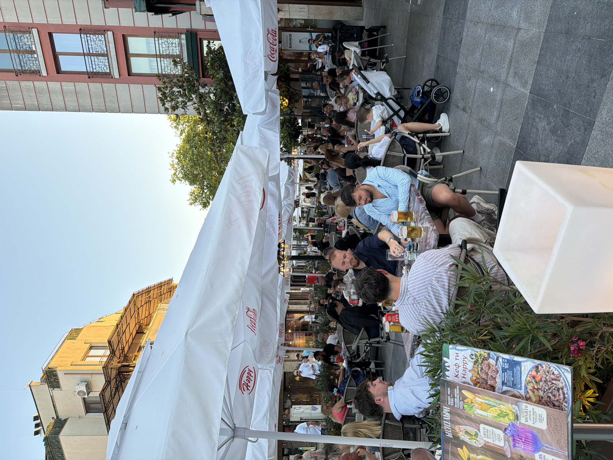 The image depicts an outdoor dining area of a restaurant in Burgas, Bulgaria. The scene is bustling with activity as patrons are seated at tables under large white umbrellas, enjoying meals and drinks. Several people are visible, including couples, families with children, and groups of friends. The architecture of the surrounding buildings is evident, with one structure featuring a distinctive salmon-colored facade and balconies with intricate metal railings, while another is a lighter-colored building with solar panels on the roof. The lighting suggests it is late afternoon or early evening, with a warm glow illuminating the scene. Menu boards are visible, displaying food and drink options in what appears to be Bulgarian script. The ground is paved with large square tiles. The overall atmosphere is one of leisure and social gathering in a European urban setting.