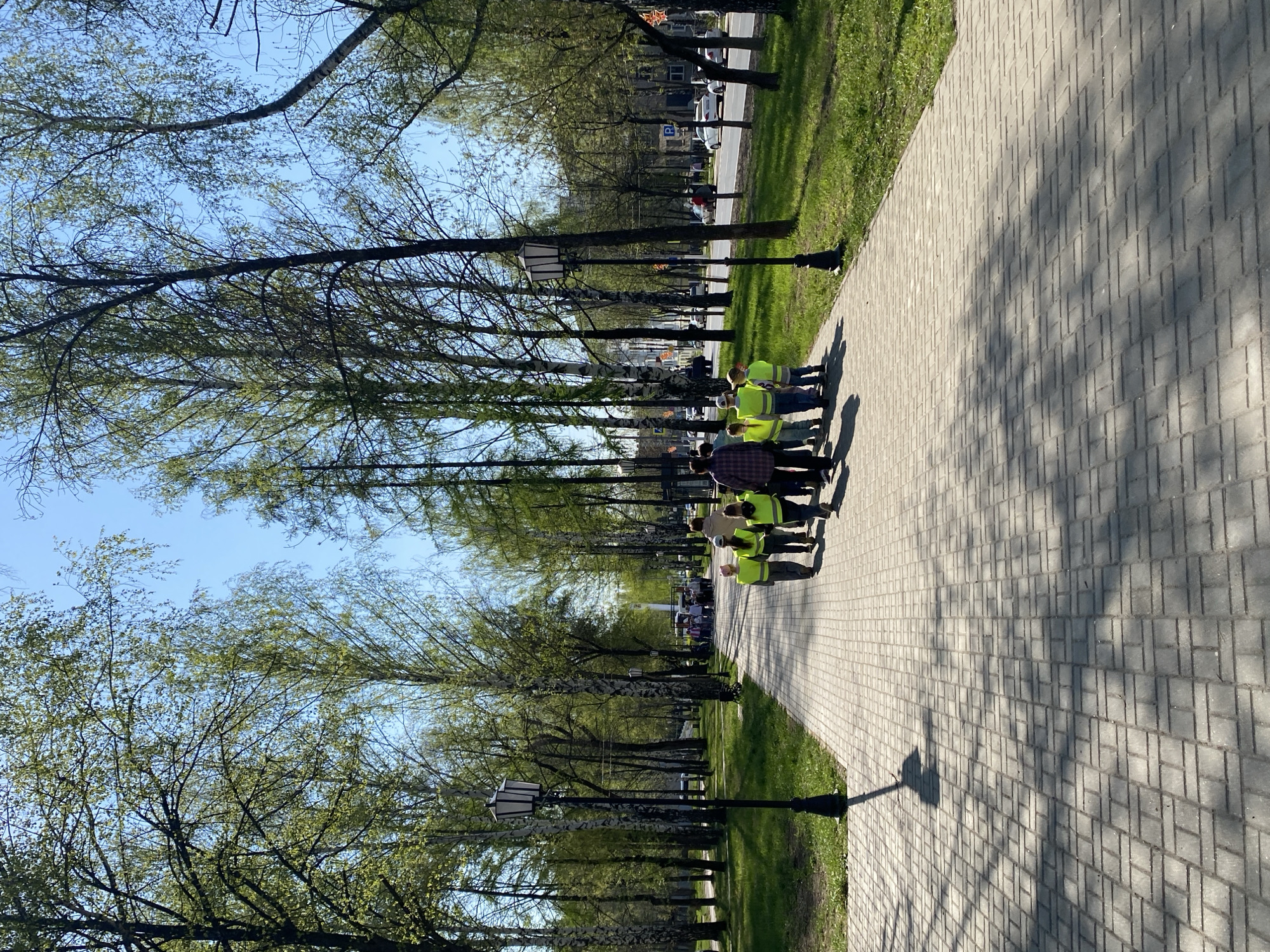 A group of people wearing yellow safety vests are walking along a brick-paved path. Trees line both sides of the path, with streetlights interspersed among them. Sunlight filters through the trees, casting shadows on the path. In the background, buildings and a street with vehicles can be seen. This location appears to be a park or pedestrian area in Chaikovsky, Russia.