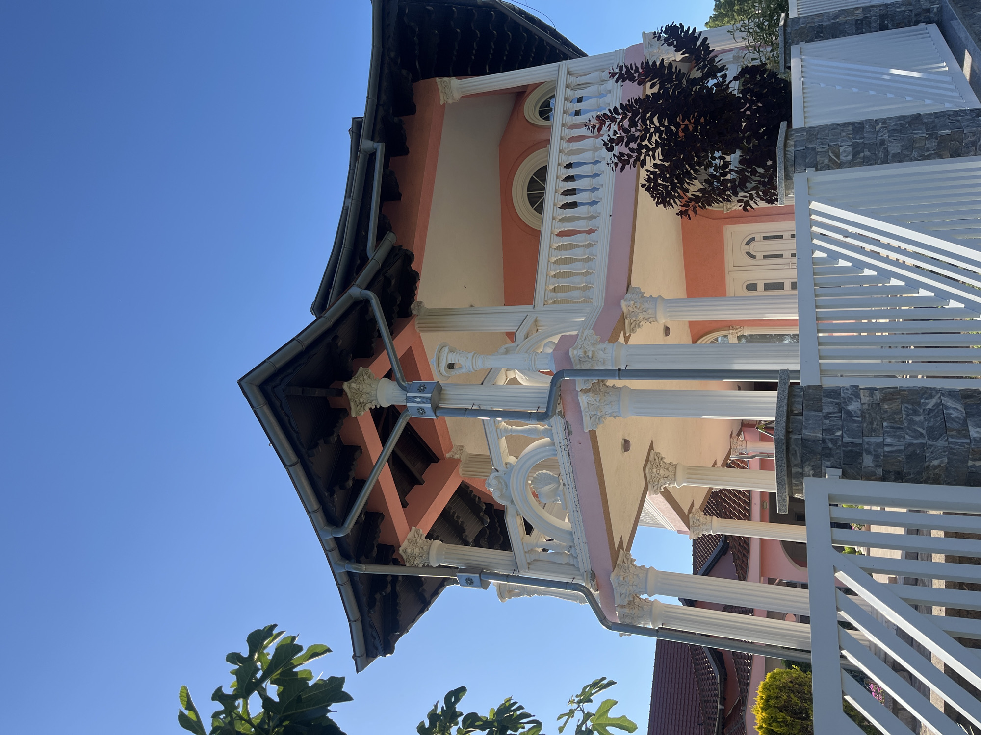 The image captures a large, multi-story house with distinct architectural features, situated in Petrovac-na-Mlavi Municipality, Serbia. The house is painted a vibrant pink and features a prominent balcony supported by numerous white Corinthian columns. The balcony is adorned with an ornate white railing. The roof is dark and appears to be tiled, with visible gutters. Two arched windows are visible on the upper level. In the foreground, a white picket fence with stone-capped pillars separates the property from the street. A dark-leaved shrub is planted in front of the house, partially obscuring the ground floor. To the left of the house, another building with a red-tiled roof is partially visible, as well as a small green shrub. The sky is a clear, bright blue. The scene is bathed in sunlight, suggesting a clear day. No people or animals are visible in the image.