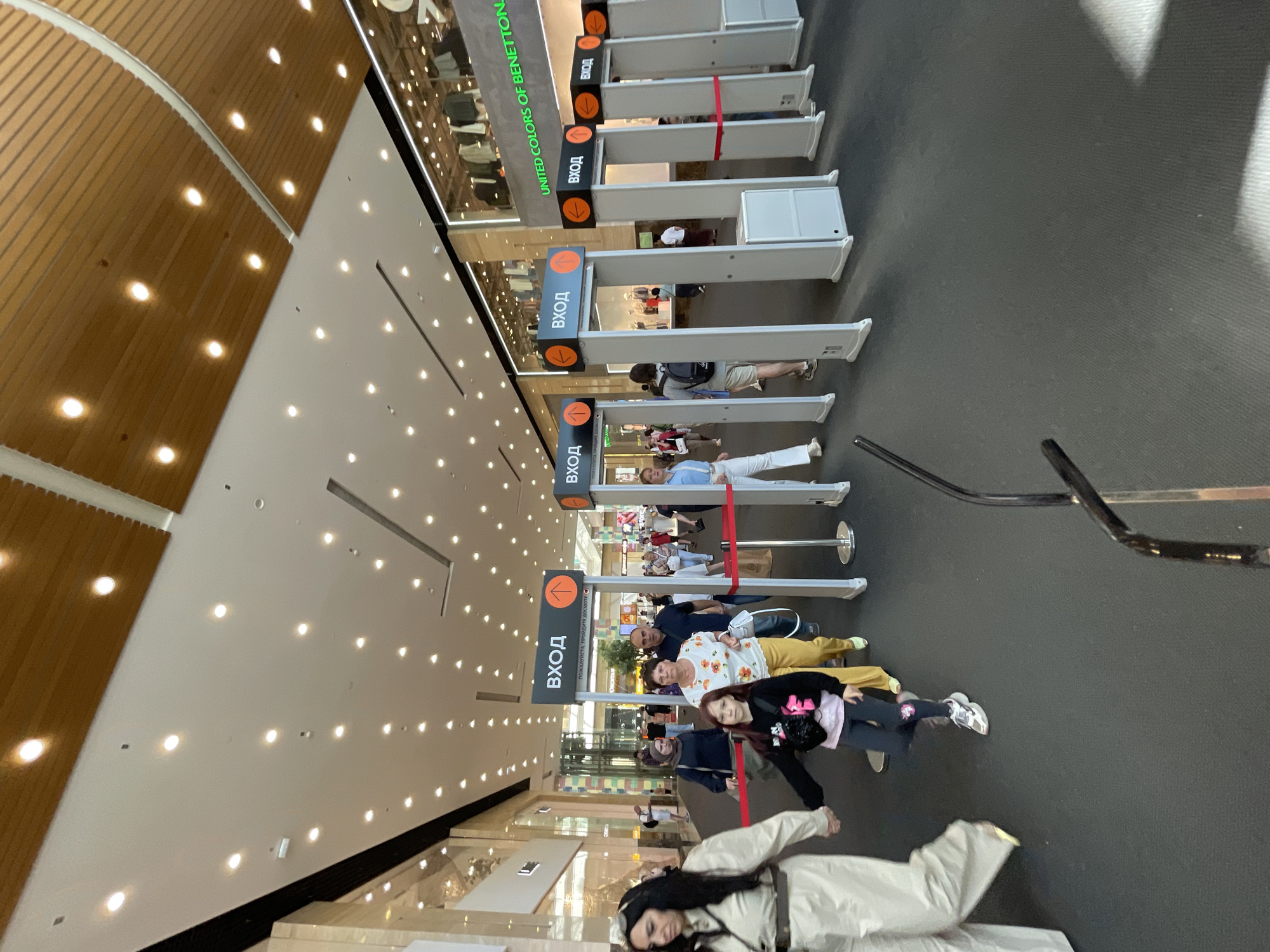 The image depicts the entrance area of a modern shopping mall in Moscow, Russia. The scene is brightly illuminated by numerous recessed circular lights embedded in a white ceiling, which also features sections of light-toned wooden slatted panels. The floor is covered with a dark gray carpet.
In the mid-ground, a series of at least seven security archways (metal detectors) are arranged in a row, serving as entry points into the mall. Each archway has an overhead sign displaying the Russian word "ВХОД" (ENTRANCE) in white text on a dark gray background, accompanied by a circular orange emblem containing an upward-pointing arrow. Below the "ВХОД" sign on the leftmost visible archway, additional Russian text reads "ПОЖАЛУЙСТА, ПРОЙДИТЕ ДОСМОТР!" (PLEASE PROCEED TO INSPECTION!). Red retractable barriers are partially visible, directing pedestrian flow between some of the archways.
Numerous individuals are observed entering the mall through these security gates. In the foreground on the left, a woman with dark hair, wearing a light-colored (possibly beige or khaki) jumpsuit and carrying a light-colored shoulder bag, walks towards the left while holding the right hand of a young girl. The girl wears a black hoodie with a pink design, dark leggings with a small colorful patch on the right knee, and gray and white sneakers. To their right, a woman in a white t-shirt with a floral pattern and yellow pants is walking, carrying a white bag. Behind her, a man in a dark polo shirt is also entering. Further into the frame, a woman wearing a dark head covering (hijab) and a dark top is visible. On the right side of the frame, several other people are passing through the archways, including a person with a dark backpack and a woman in a light blue shirt and white pants.
In the background, storefronts are visible, including one prominently featuring "UNITED COLORS OF BENETTON" in green text, indicating a retail presence within the mall. Other retail displays are visible through large glass windows. The overall impression is one of a busy commercial environment with security measures in place for entry.