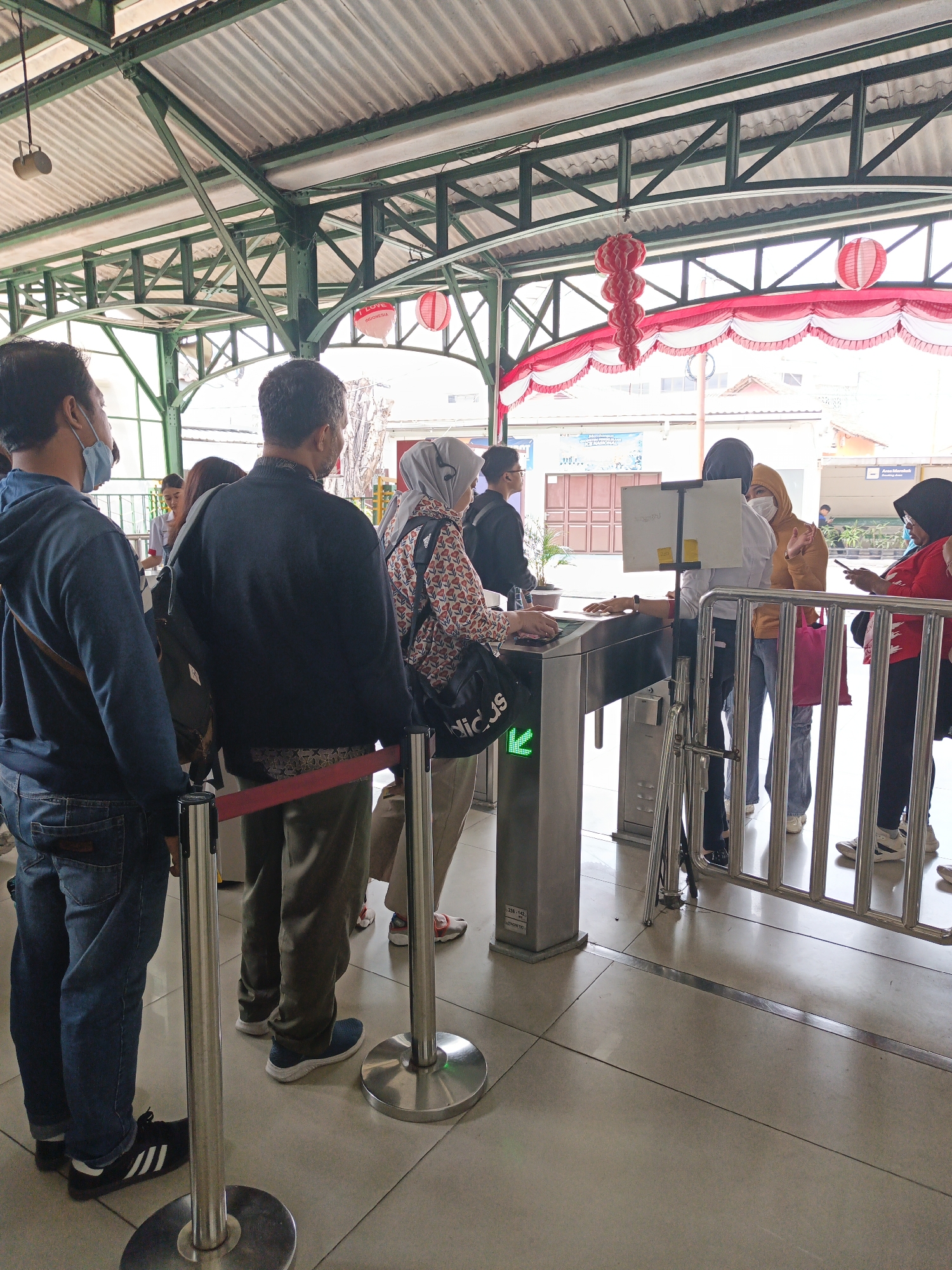 The image depicts a group of people queuing at a ticket gate, likely at a public transportation hub such as a train station or bus terminal. The location is identified as Surakarta, Indonesia, indicated by subtle contextual clues such as the architectural style and the presence of red and white decorative elements, which are common during national celebrations in Indonesia.
In the foreground, several individuals are visible, facing away from the camera and towards the ticket gate. The person on the far left, wearing a blue jacket and jeans, is looking towards the gate. Next to him, a person in a dark jacket and patterned pants is also facing the gate. Further down the line, other individuals, including women wearing headscarves, are also in the queue.
The ticket gate itself is a modern, automated turnstile with a screen and card reader. A green arrow is illuminated on the gate, suggesting it is operational and allowing passage. The environment appears to be an indoor or semi-outdoor space, with a corrugated metal roof supported by green-painted metal beams. Red lanterns are hanging from the ceiling, adding a festive or decorative touch. The floor is tiled. The overall scene conveys a sense of order and routine as people wait to pass through the gate.