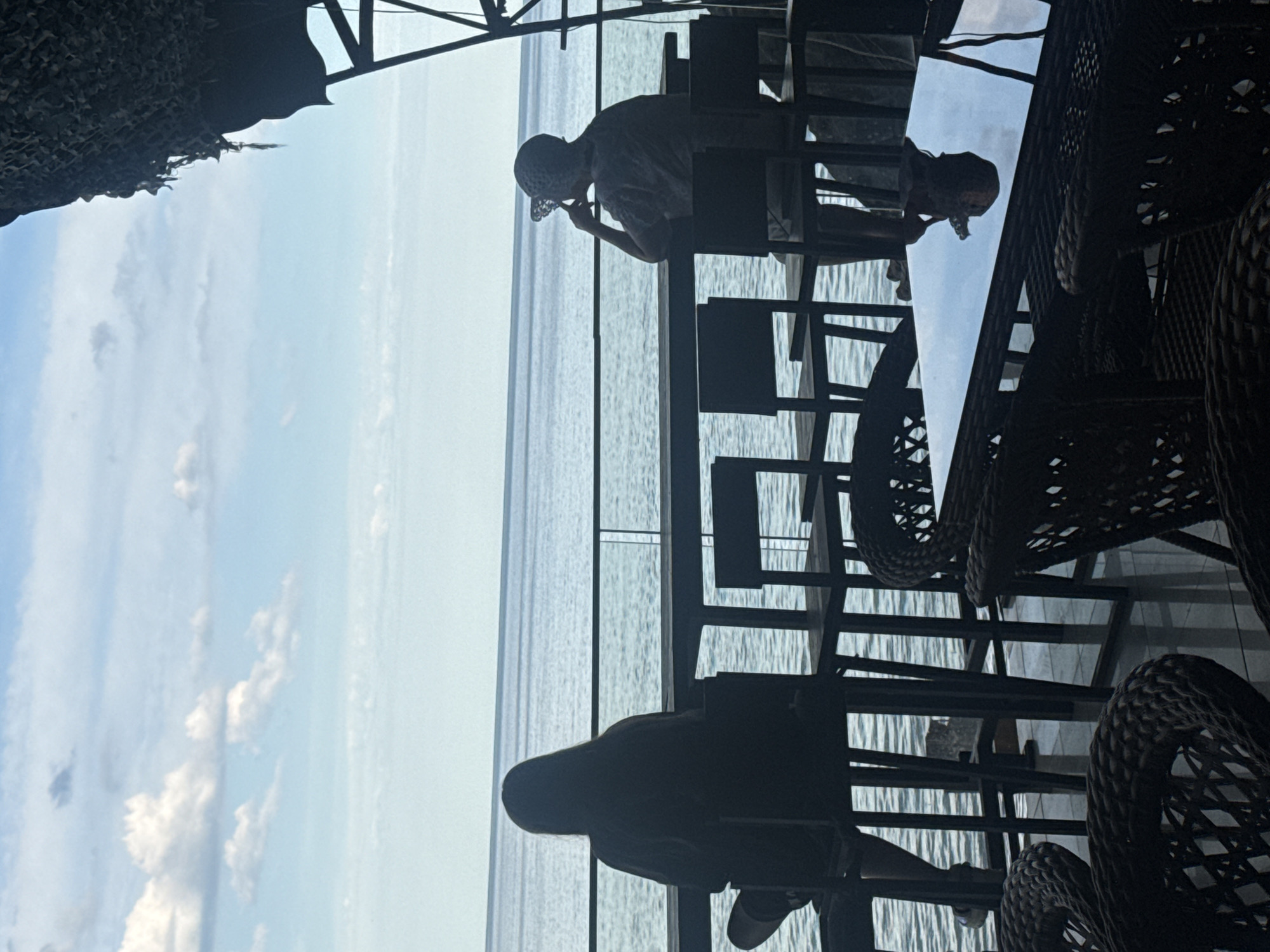 Two people are seated at a high table overlooking a body of water. The person on the left, facing away from the camera, has long dark hair and is wearing a dark shirt. The person on the right, also facing away from the camera, is wearing a hat and a patterned shirt, with their hand resting on their chin. They are both seated on high chairs. In the foreground, rattan chairs and a glass-top table are visible, reflecting the sky and clouds. The background shows a calm, blue body of water meeting a pale blue sky with scattered white clouds. A portion of a camouflage-like canopy structure is present in the upper right corner of the image. The scene appears to be an outdoor seating area of a restaurant or cafe.