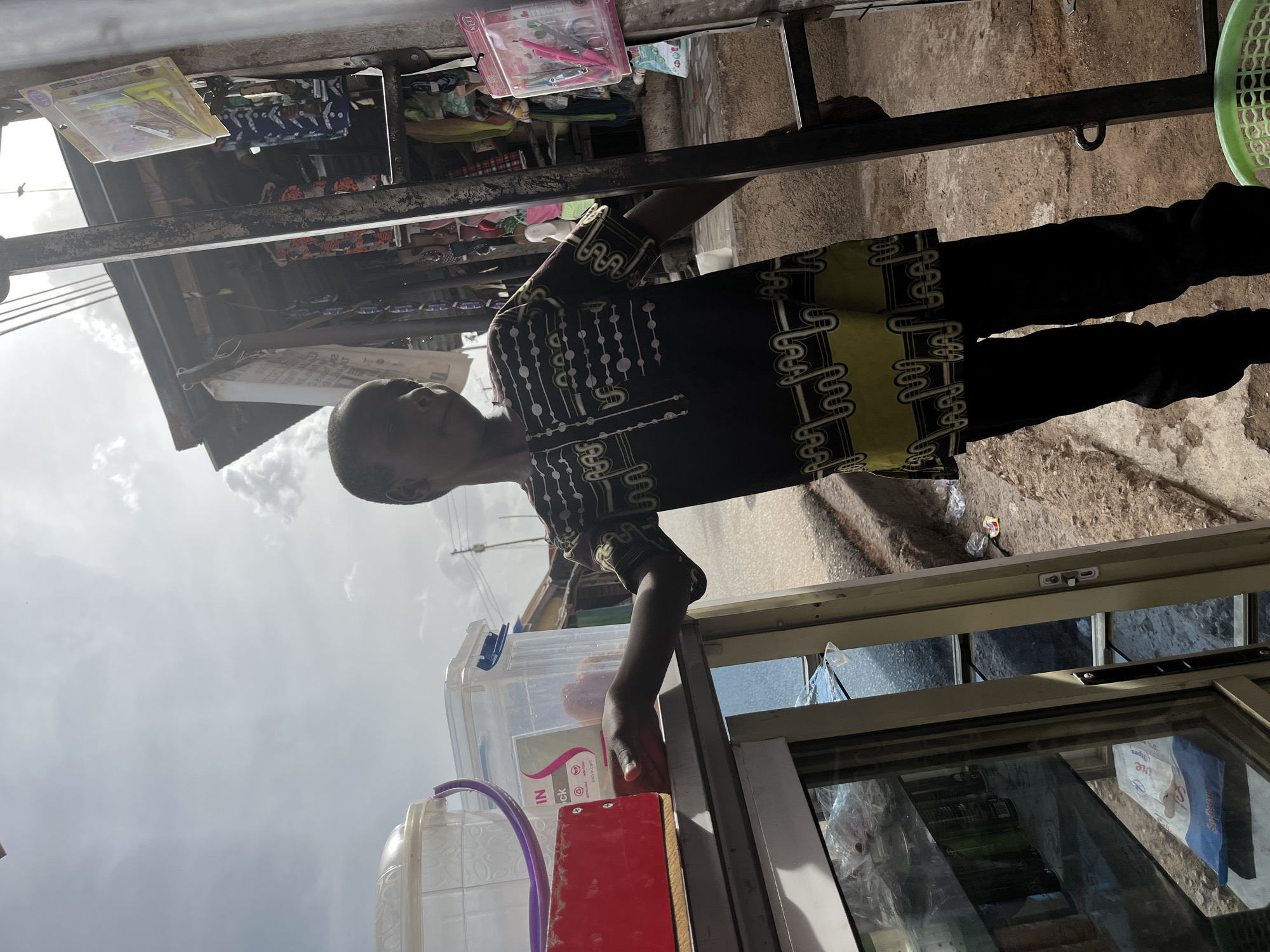 A child stands next to a glass-fronted kiosk in Jos, Nigeria. The child wears a dark, patterned shirt and dark pants. Plastic containers and a red object sit atop the kiosk. Merchandise is displayed above the kiosk, under a metal awning. The background shows a street scene, and the sky is visible.