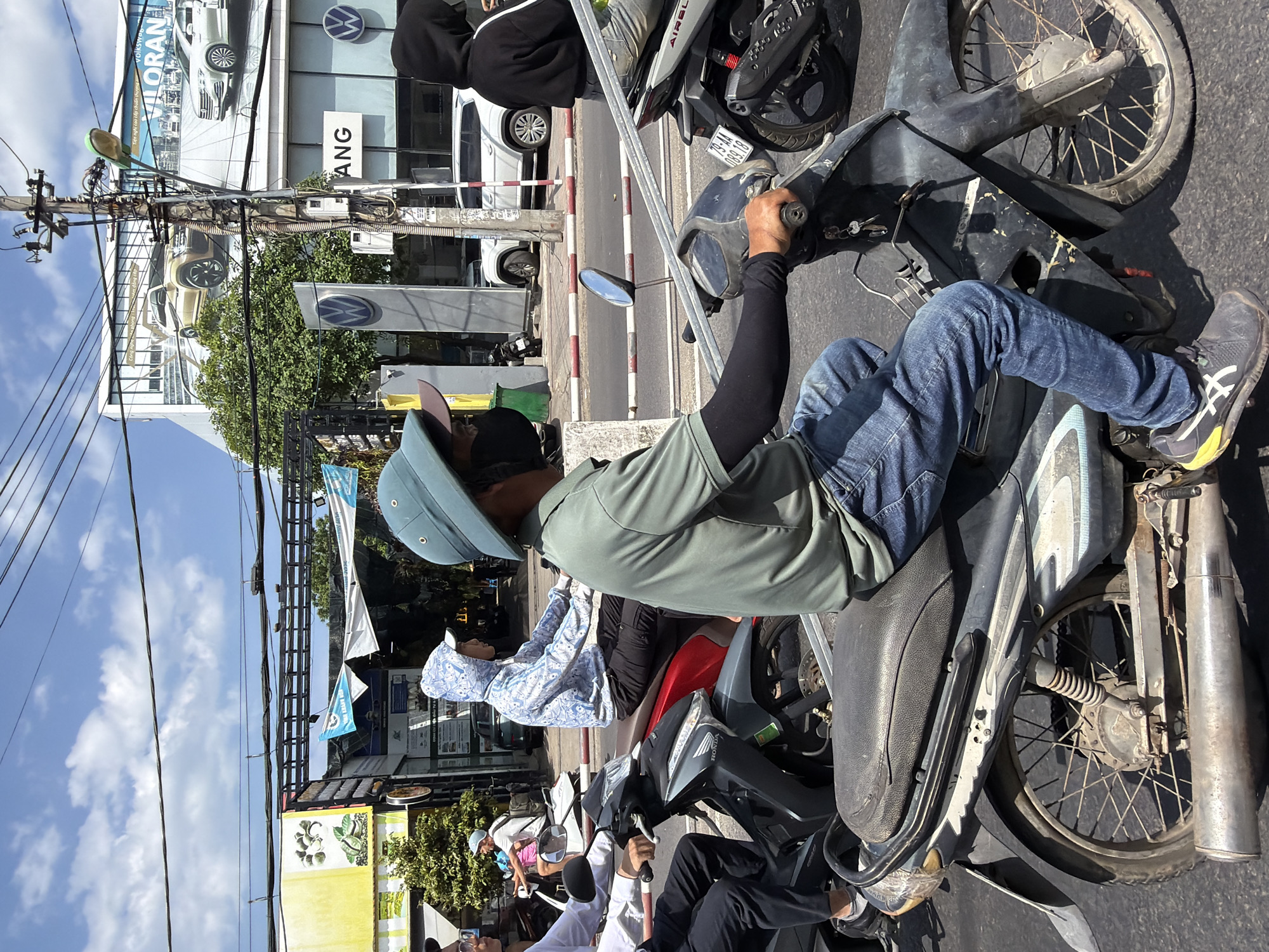 This image captures a street scene in Nha Trang, Vietnam, likely on a sunny day, given the bright lighting and shadows. The foreground is dominated by a dark-colored motorcycle, on which a person wearing a green shirt, blue jeans, and a dark face mask is seated. This individual is also wearing a wide-brimmed hat, which is a common accessory in sunny climates. Their right hand is on the handlebar of the motorcycle. Behind this person, another motorcycle is visible, with a rider wearing a light-colored hooded sweatshirt with a distinctive pattern and a blue baseball cap. In the background, there are several buildings, one of which appears to be a car dealership with large billboards displaying vehicles and text, possibly in Vietnamese. One billboard prominently features the Volkswagen logo and imagery of SUVs. Power lines crisscross the sky, indicating an urban environment. Other vehicles and pedestrians are present in the scene, suggesting a busy street. The road surface is asphalt, with painted lines and a red and white striped barrier visible in the middle ground. The overall impression is one of everyday life and traffic in a Vietnamese city.