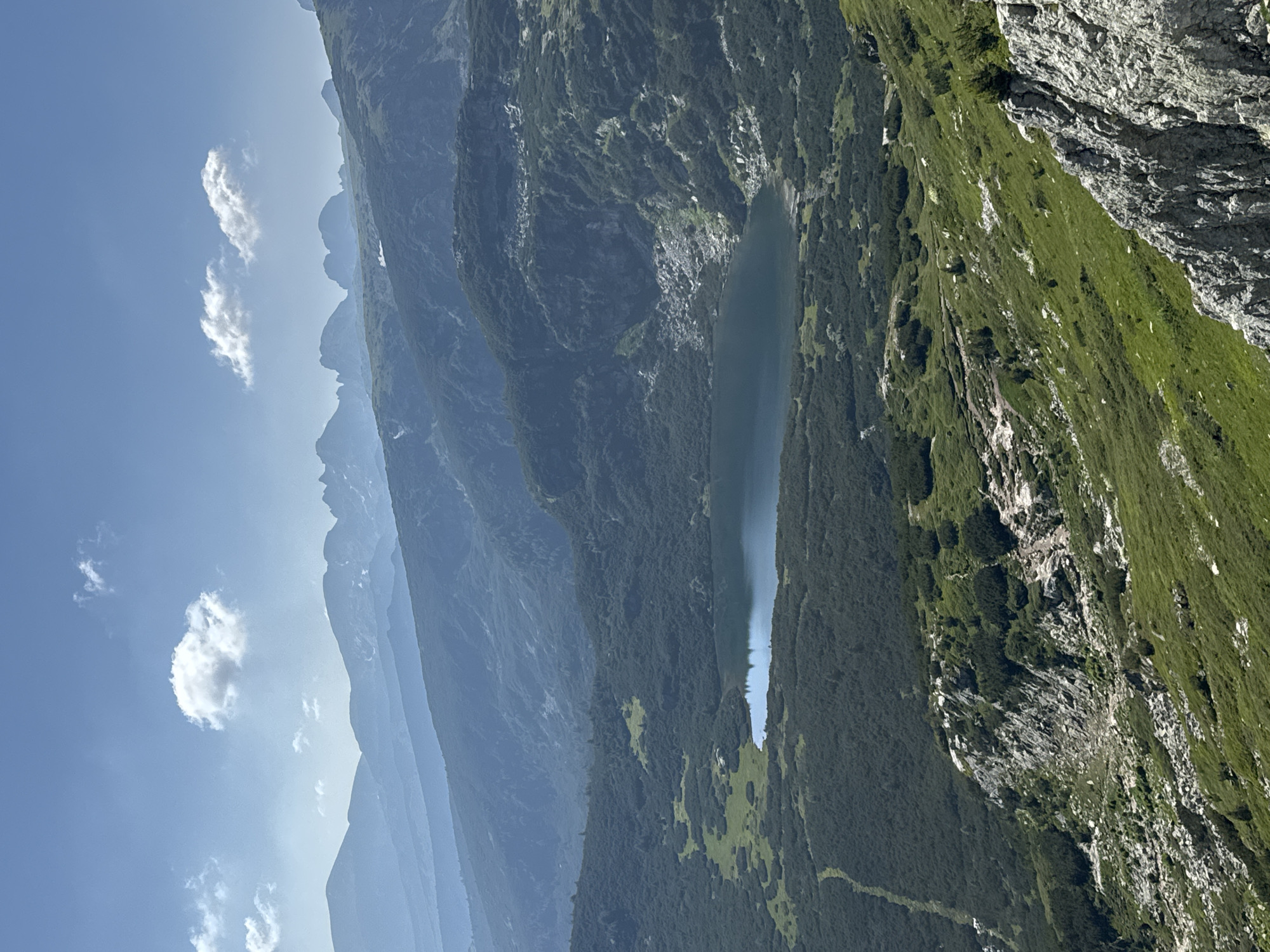 A high-angle, wide shot captures a vast mountain landscape on a clear, sunny day. In the foreground, a steep, rocky slope covered in green grass and sparse shrubs descends from the bottom right. The middle ground is dominated by a tranquil, dark blue lake nestled within a valley. Surrounding the lake is dense, dark green coniferous forest. Beyond the valley, successive layers of mountains, fading into a hazy blue in the distance, stretch across the horizon under a bright blue sky with a few scattered white clouds. The scene is located in the Rila Mountains, near Panichishte, Bulgaria.