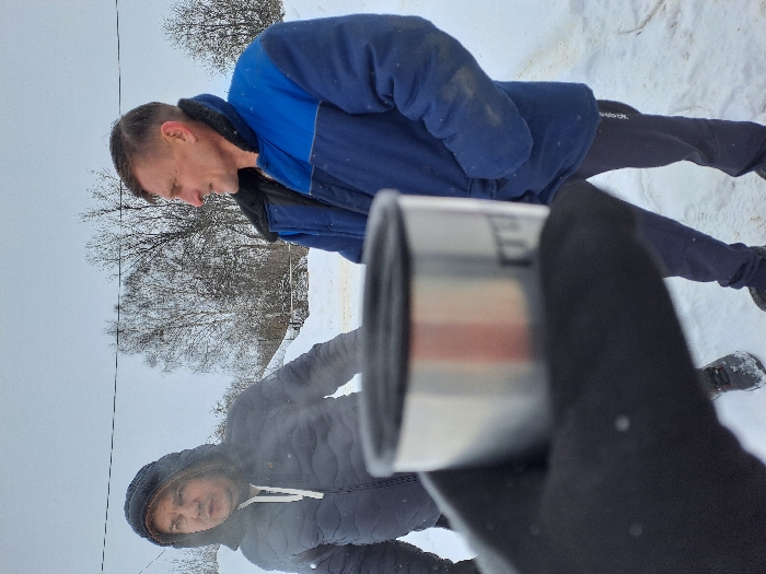 The image depicts an outdoor winter scene in the city of Полянки, Russia. The ground is covered in undisturbed snow, and bare trees are visible in the background under an overcast sky. In the foreground, a gloved hand holds a silver-colored metallic mug with a black rim, appearing out of focus. Two men stand in the mid-ground. The man on the left wears a dark blue or black puffy winter jacket with the hood up; he is looking towards the right with a slight opening of his mouth, suggesting speech or exhaling breath in the cold air. The man on the right wears a two-tone blue and dark blue winter work jacket with a black collar, dark trousers, and dark footwear; he has short, dark hair and is looking slightly to his left with a subtle smile, his hands in his pockets. Both individuals are dressed for cold weather conditions.